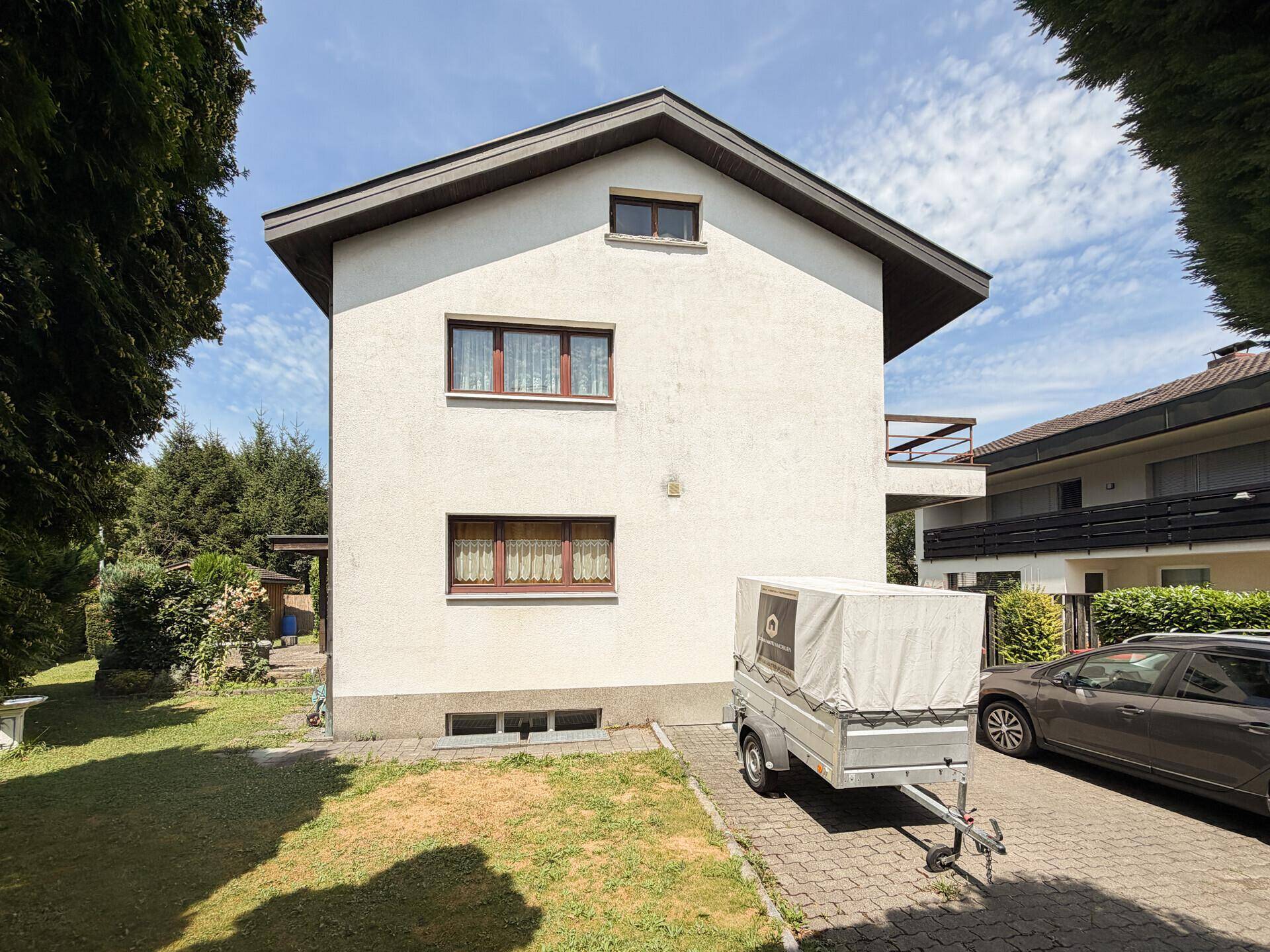 Seitenansicht des Hauses mit weißer Fassade, braunen Fensterrahmen und einem Anhänger auf dem Hof.