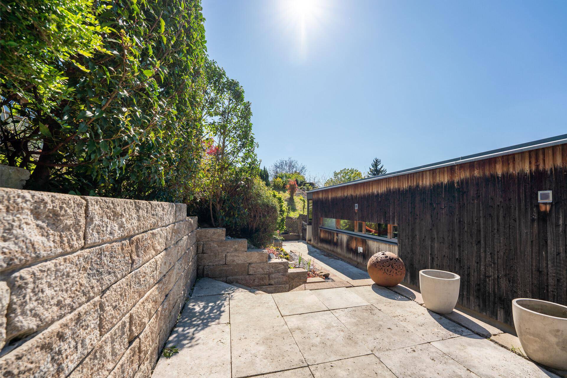 Sonnige Gartenterrasse mit Steinplatten, hohen Hecken und dekorativen Elementen.