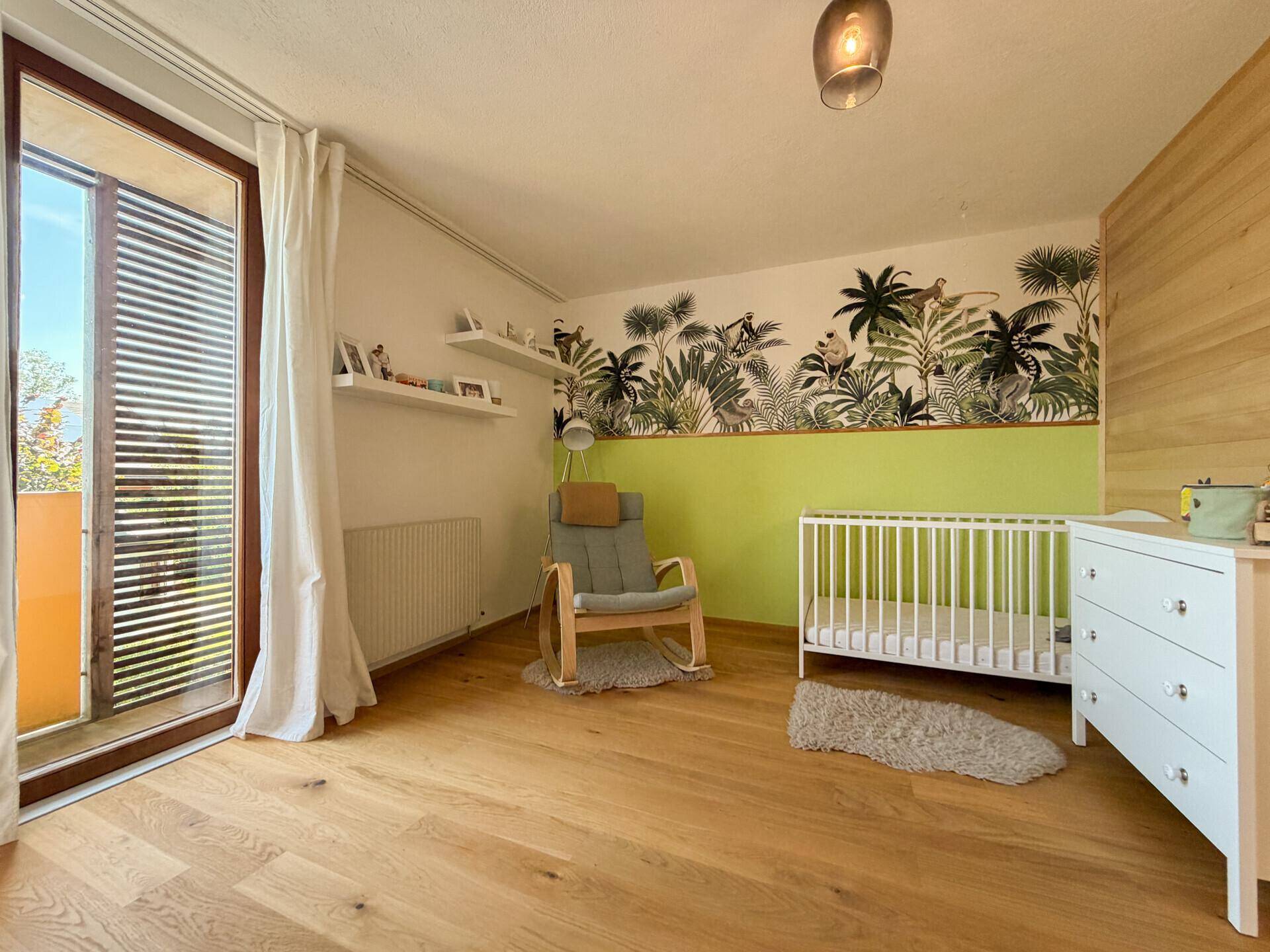 Kinderzimmer mit Holzboden, Babybett und farbenfroher Dschungel-Tapete.