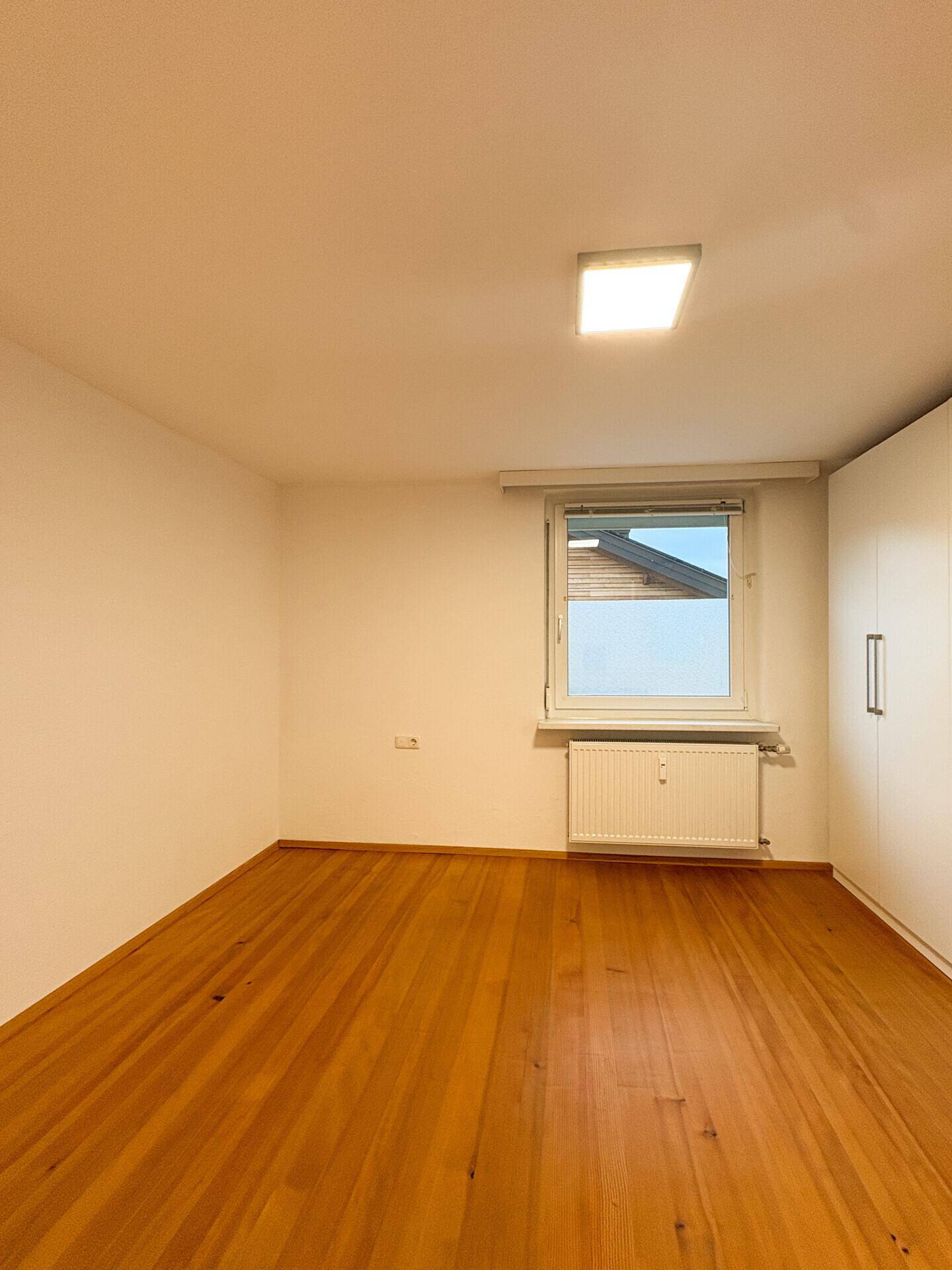 Schlafzimmer mit hellem Holzfußboden, Fenster und weißem Einbauschrank.