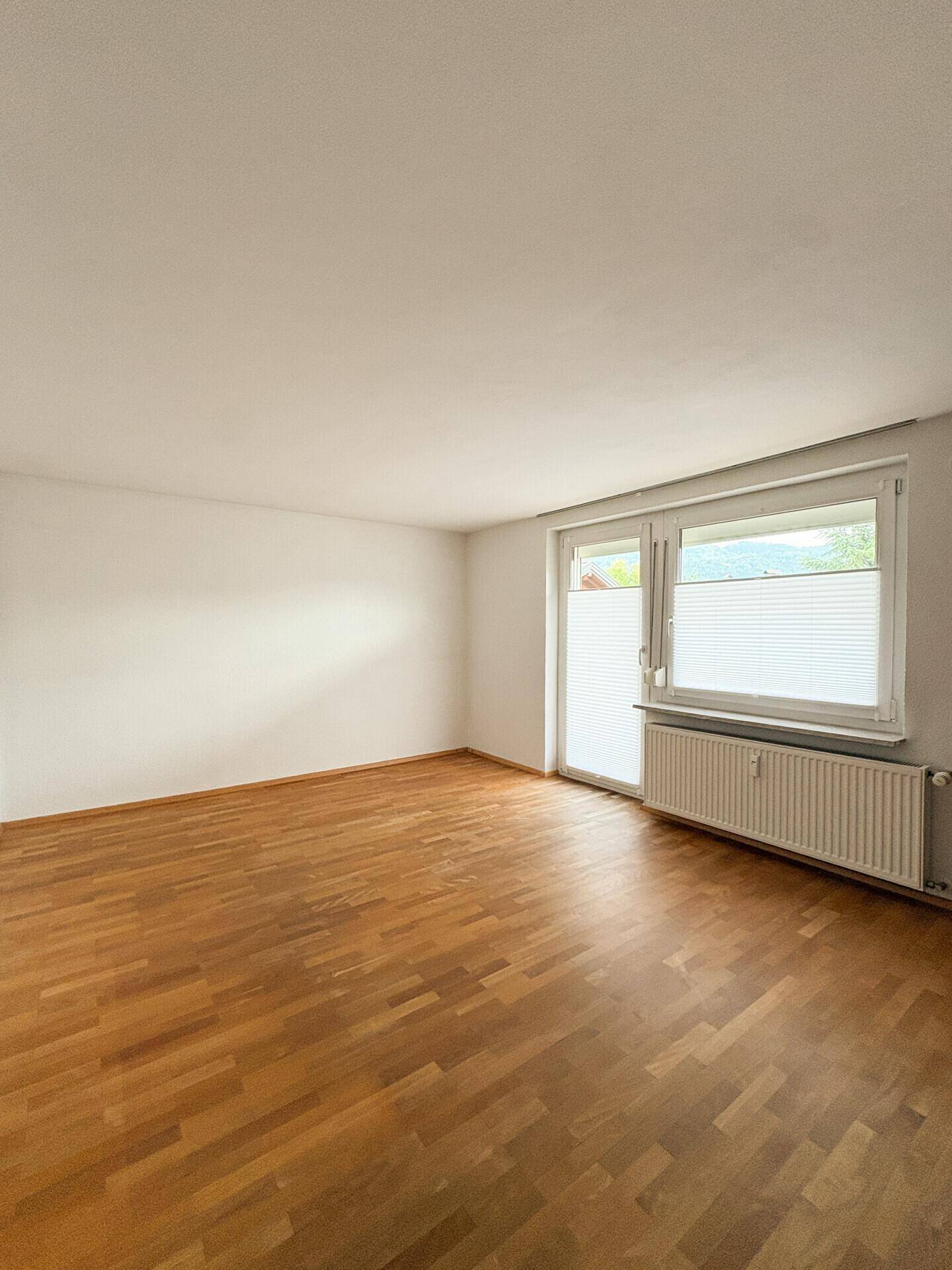 Leeres Zimmer mit hellem Parkettboden, großen Fenstern und Heizkörper.