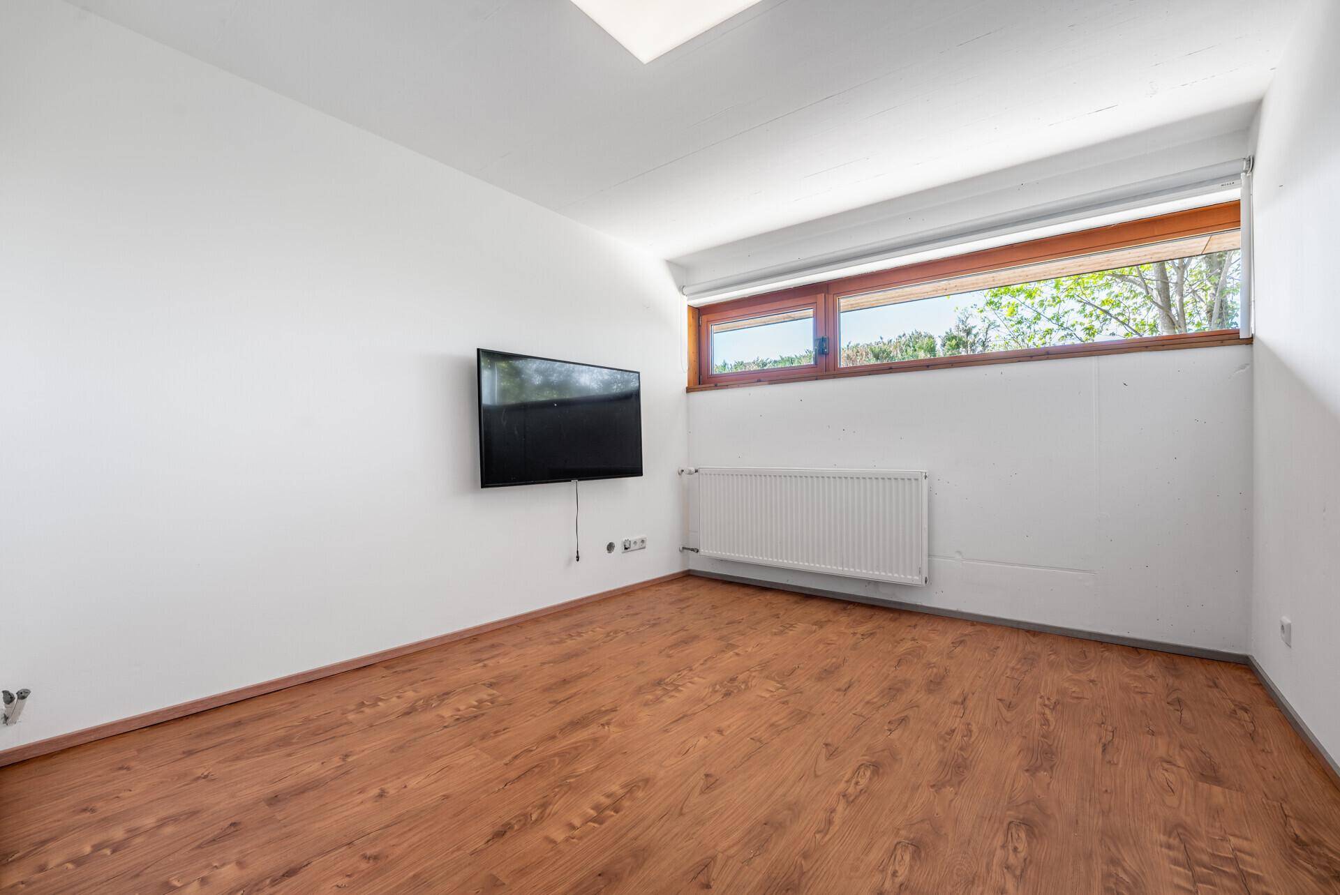 Helles Zimmer mit Holzboden, Wandfernseher und Heizkörper unter dem Fenster.