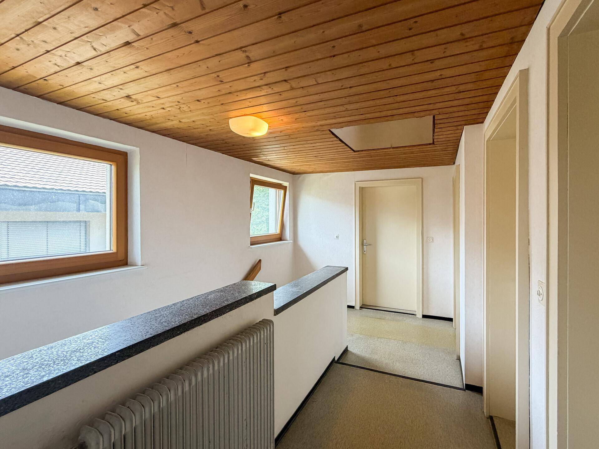 Flur im Obergeschoss mit Holzdecke, Teppichboden und Fenstern mit Blick nach draußen.