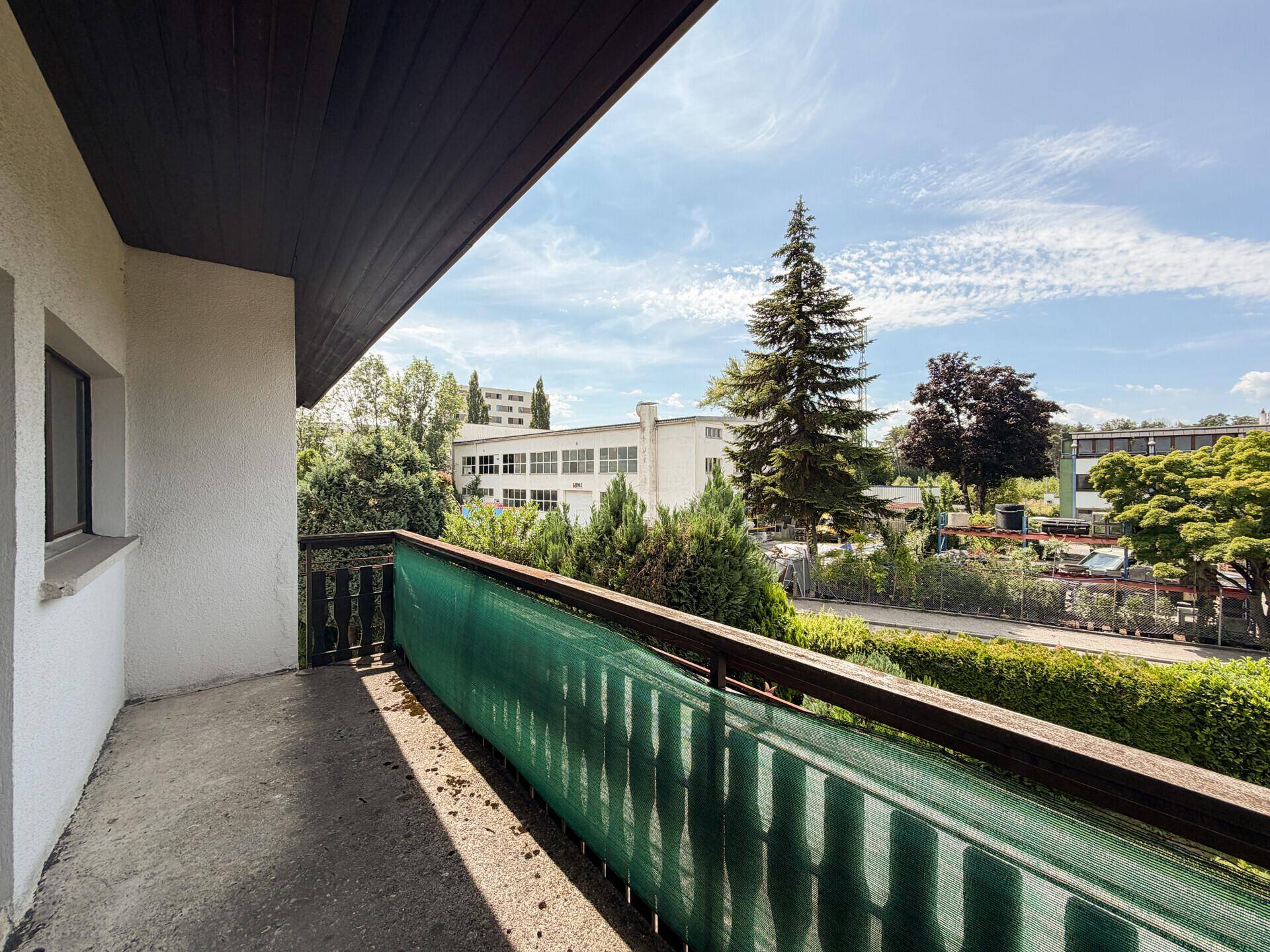 Balkon mit Holzgeländer und Blick auf die grüne Umgebung und umliegende Gebäude.