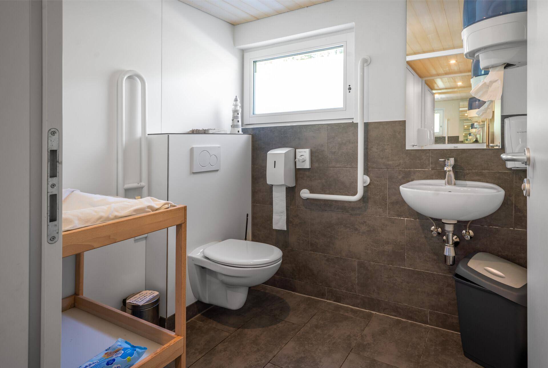 Barrierefreies Badezimmer mit Haltegriffen, Wickeltisch und Fenster, ausgestattet mit modernen Fliesen.