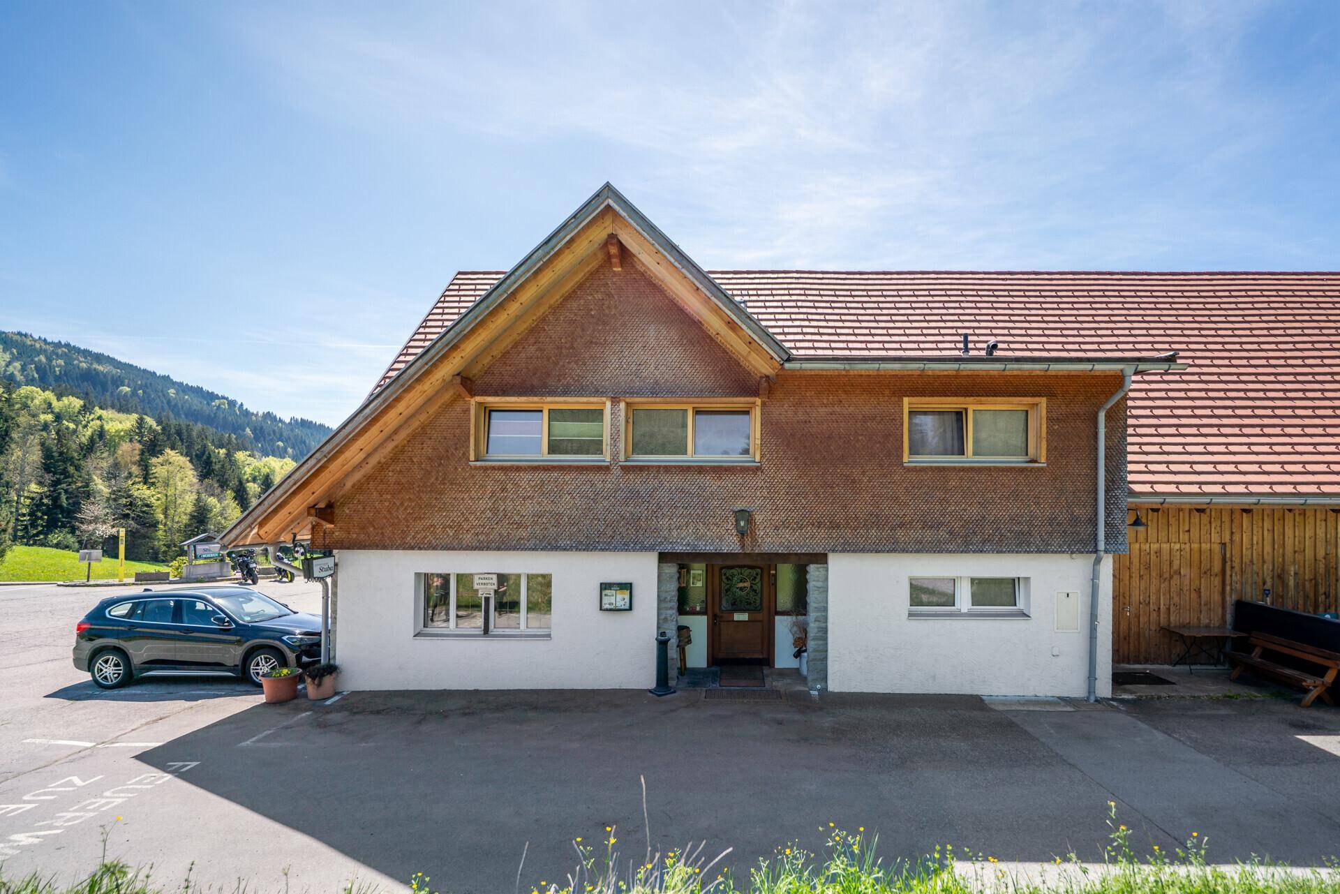 Frontansicht eines traditionellen Gebäudes mit Holzschindeln und einem Parkplatz davor, umgeben von grünen Hügeln.