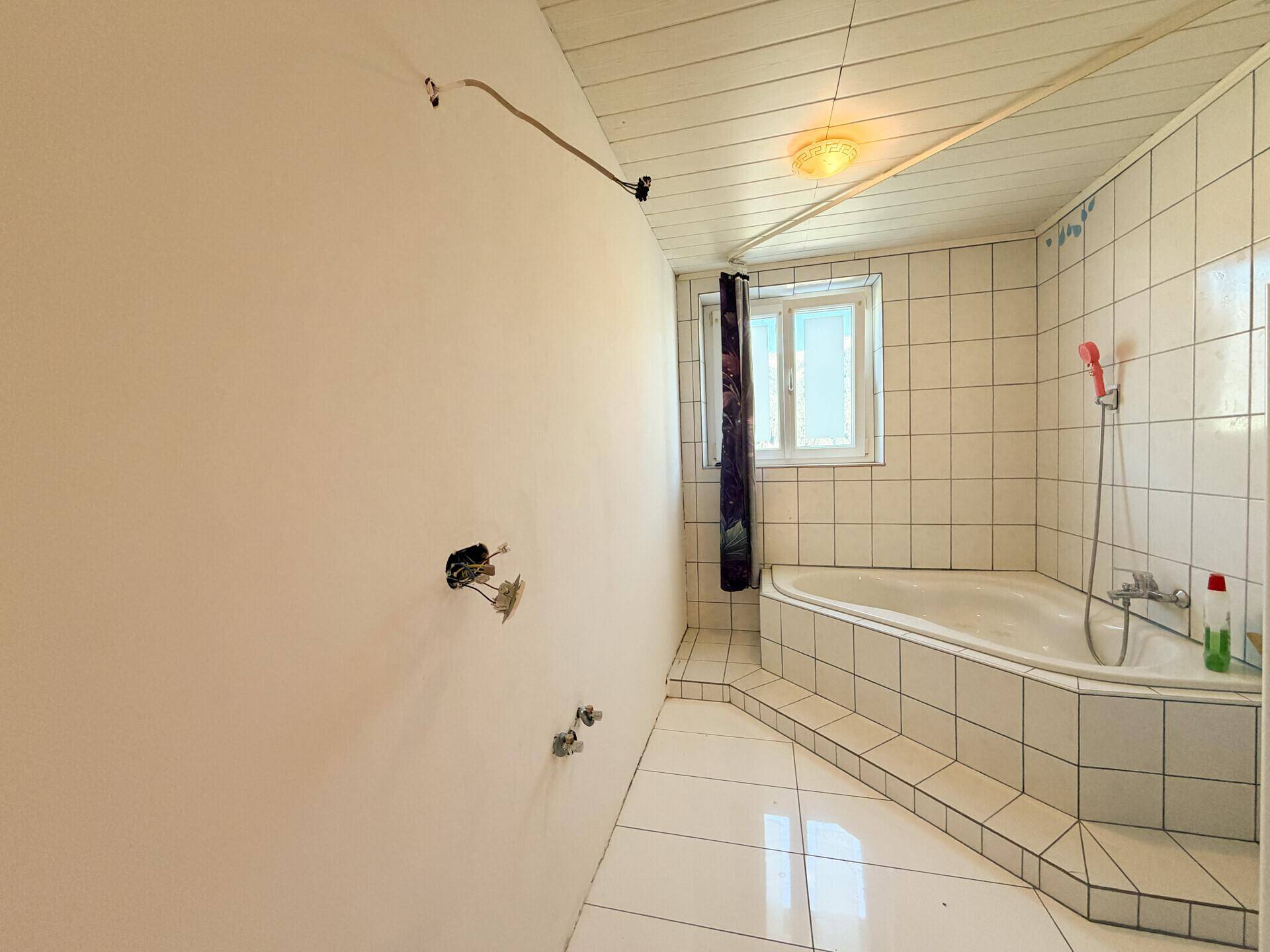 Badezimmer mit weißen Fliesen und einer Eckbadewanne, bietet ein Fenster für natürliche Belüftung.
