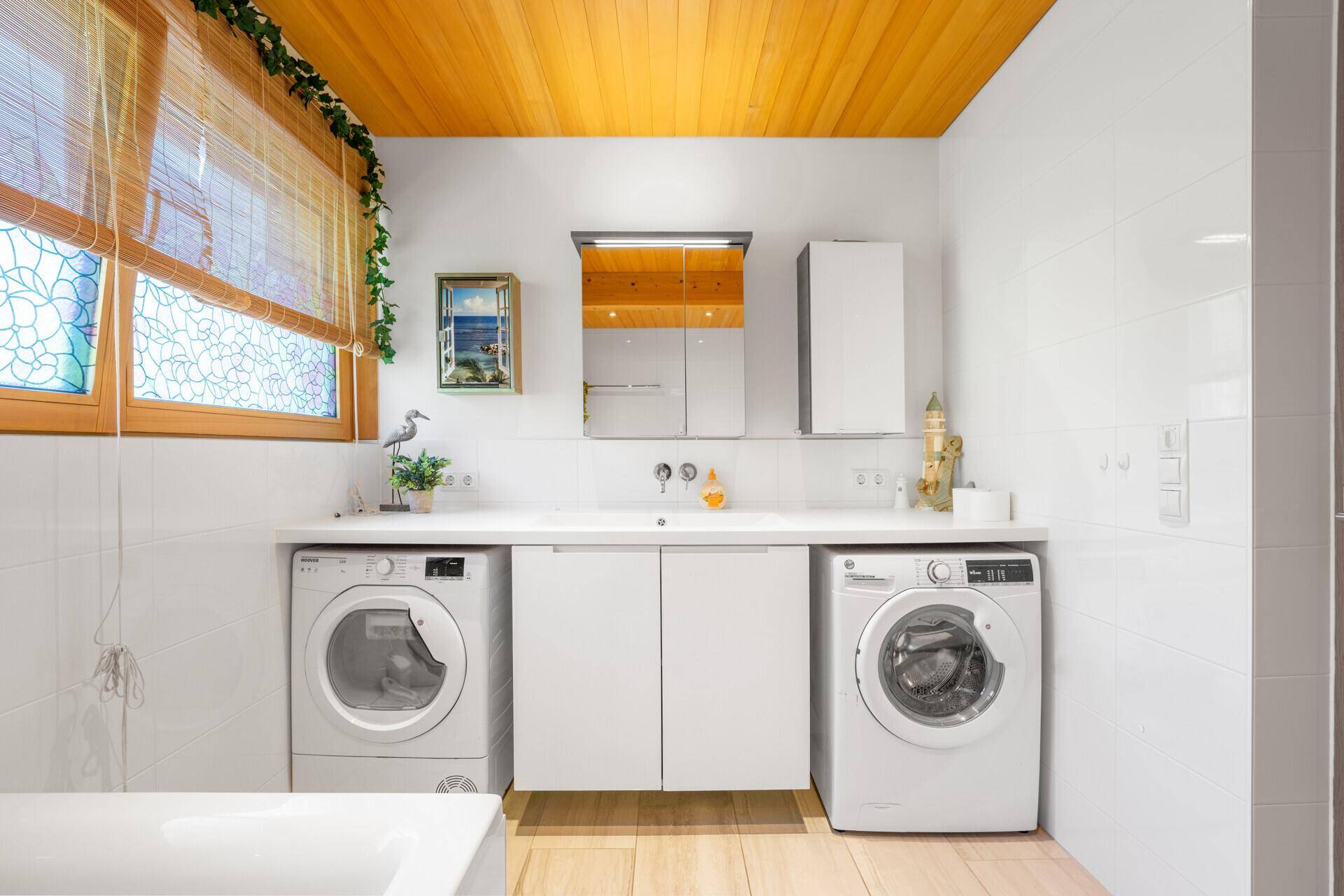 Modernes Badezimmer mit weißen Fliesenwänden, Doppelwaschtisch und zwei Waschmaschinen, mit Fenster.