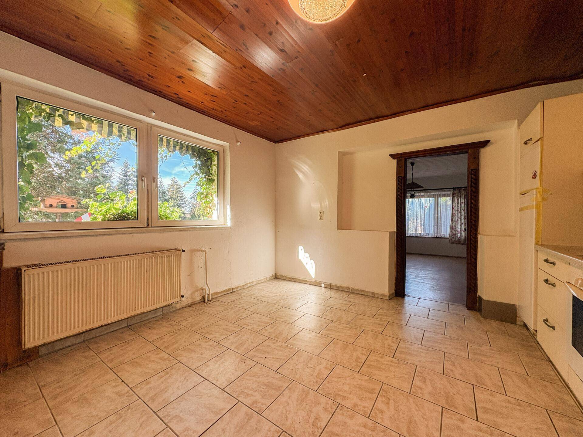 Geräumige Küche mit Fensterblick ins Grüne, Fliesenboden und Holzdecke.