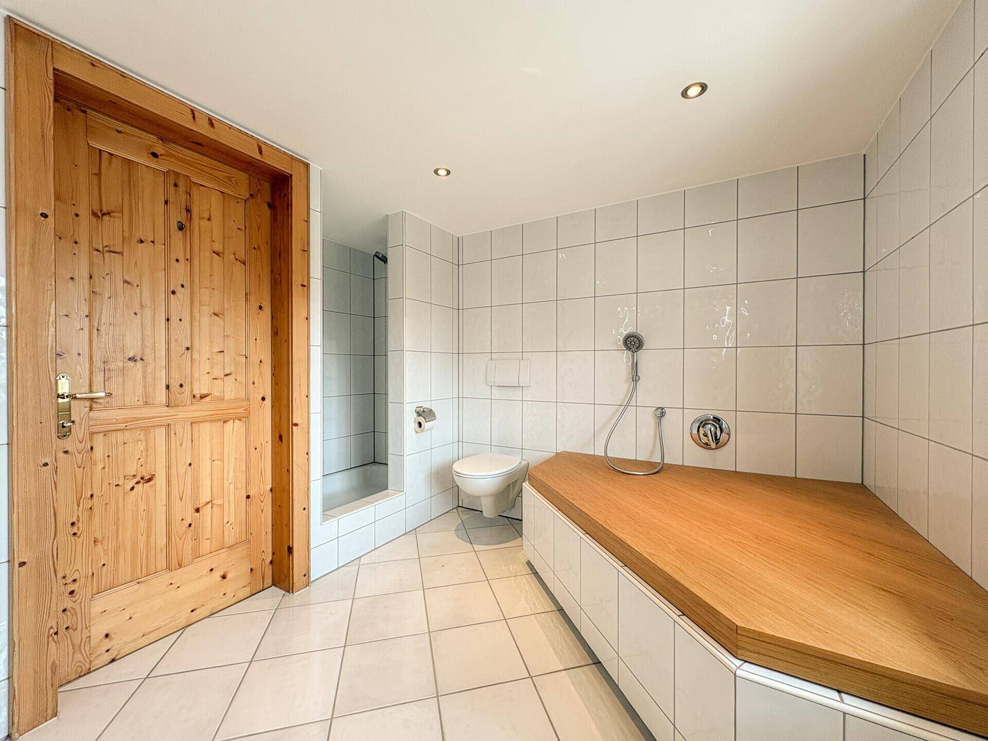Badezimmer mit weißen Fliesen, Dusche und integrierter Holzablage.