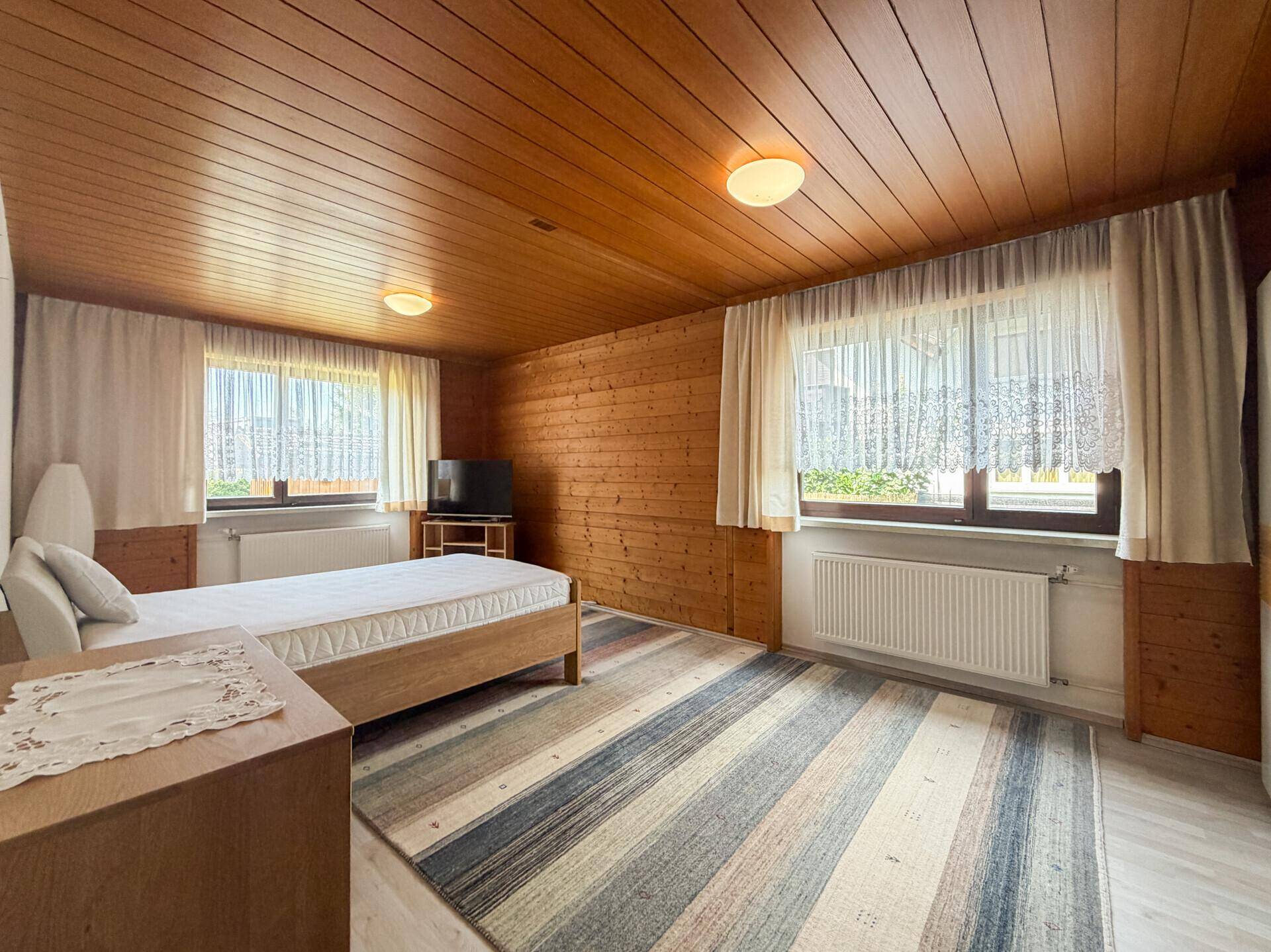 Schlafzimmer mit Holzdecke, Holzwand und Teppichboden, sowie zwei Fenstern.