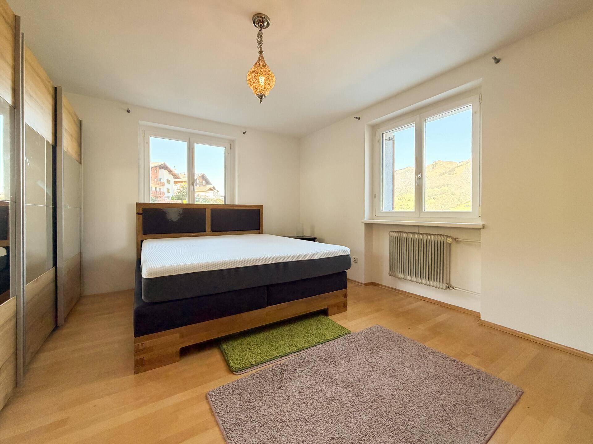 Schlafzimmer mit Holzparkettboden und einem bequemen Bett, bietet durch zwei Fenster viel Tageslicht und Bergblick.