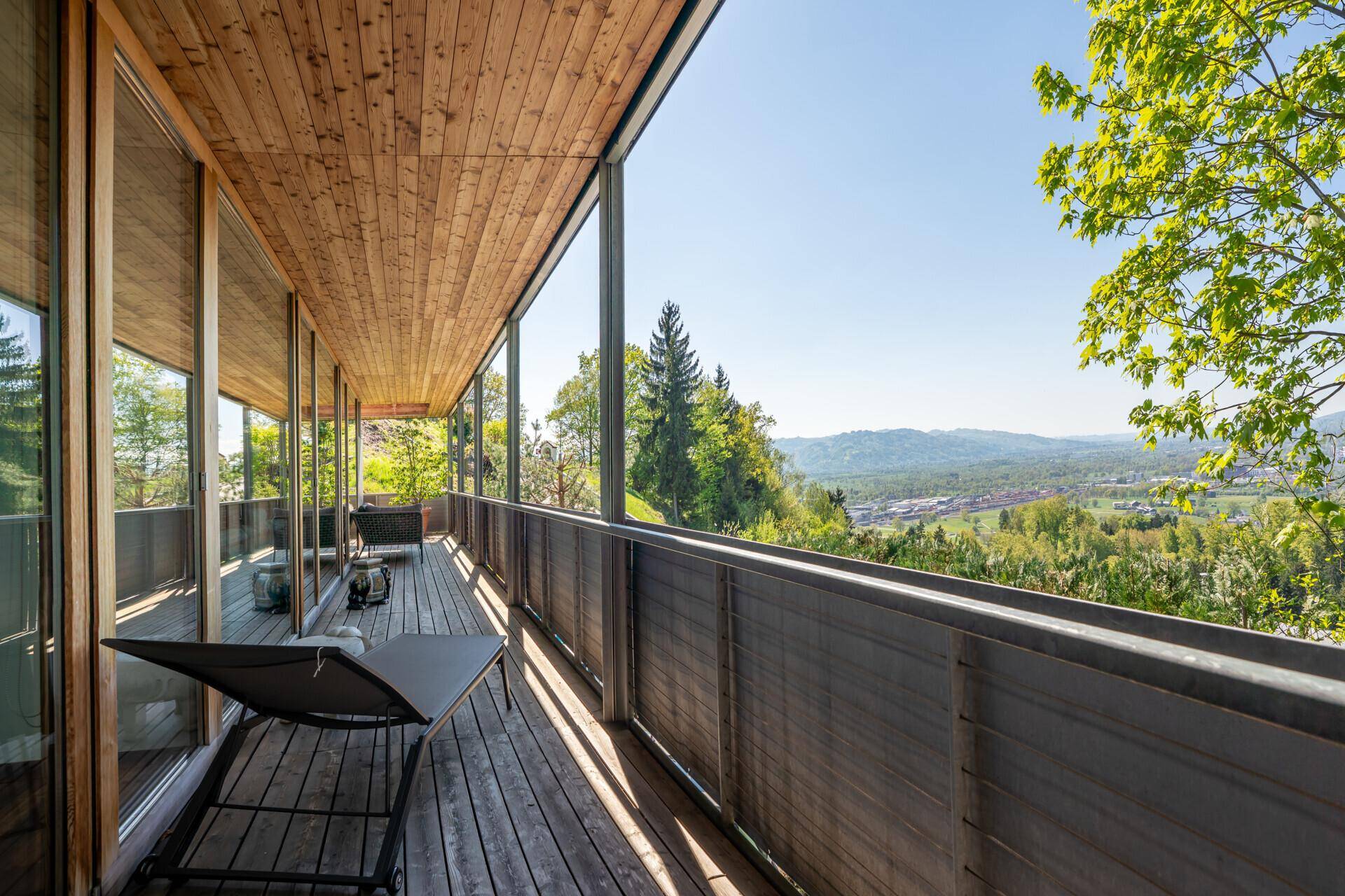 Großer Holzbalkon mit Liegestuhl und weitem Blick über die grüne Landschaft.