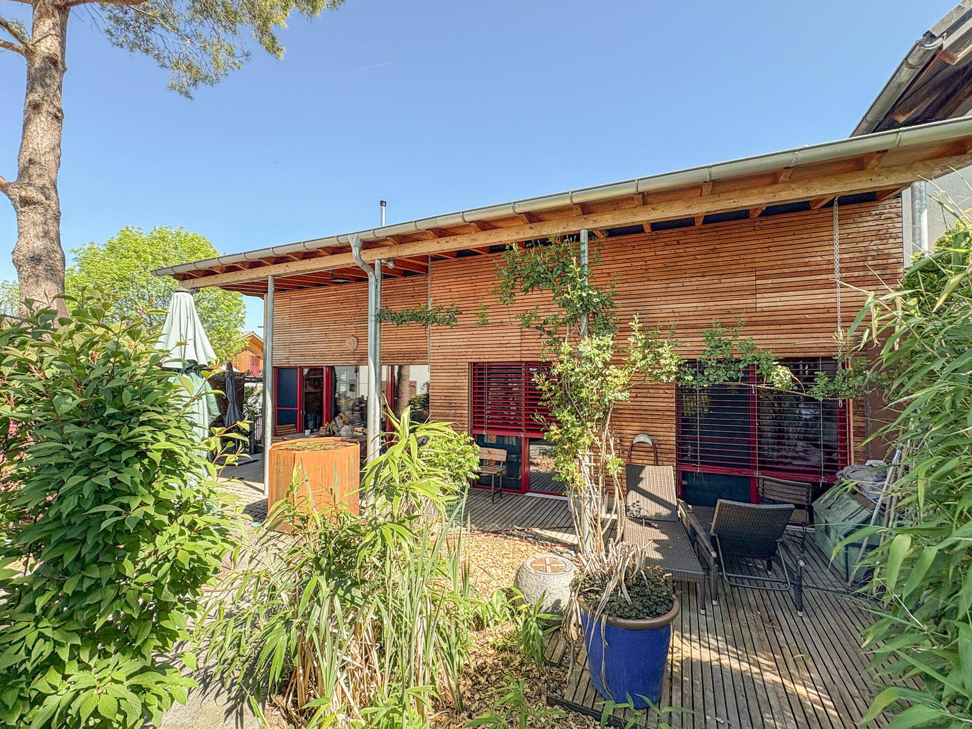 Sonnige Gartenterrasse mit Liegestühlen, üppiger Bepflanzung und Holzfassade des Hauses.