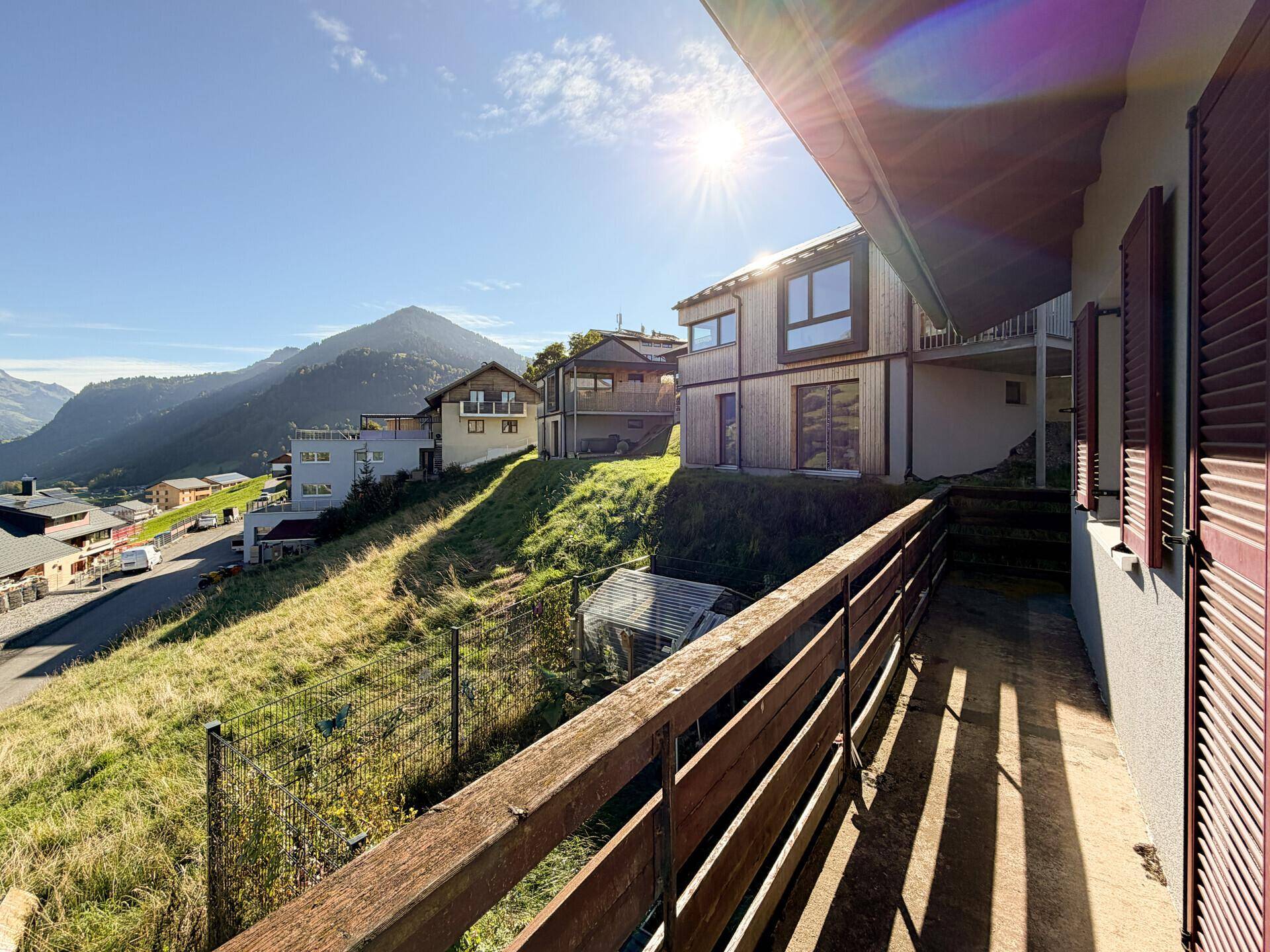 Balkon mit Holzgeländer und Blick auf die sonnige Hanglage mit weiteren Häusern und grüner Umgebung.