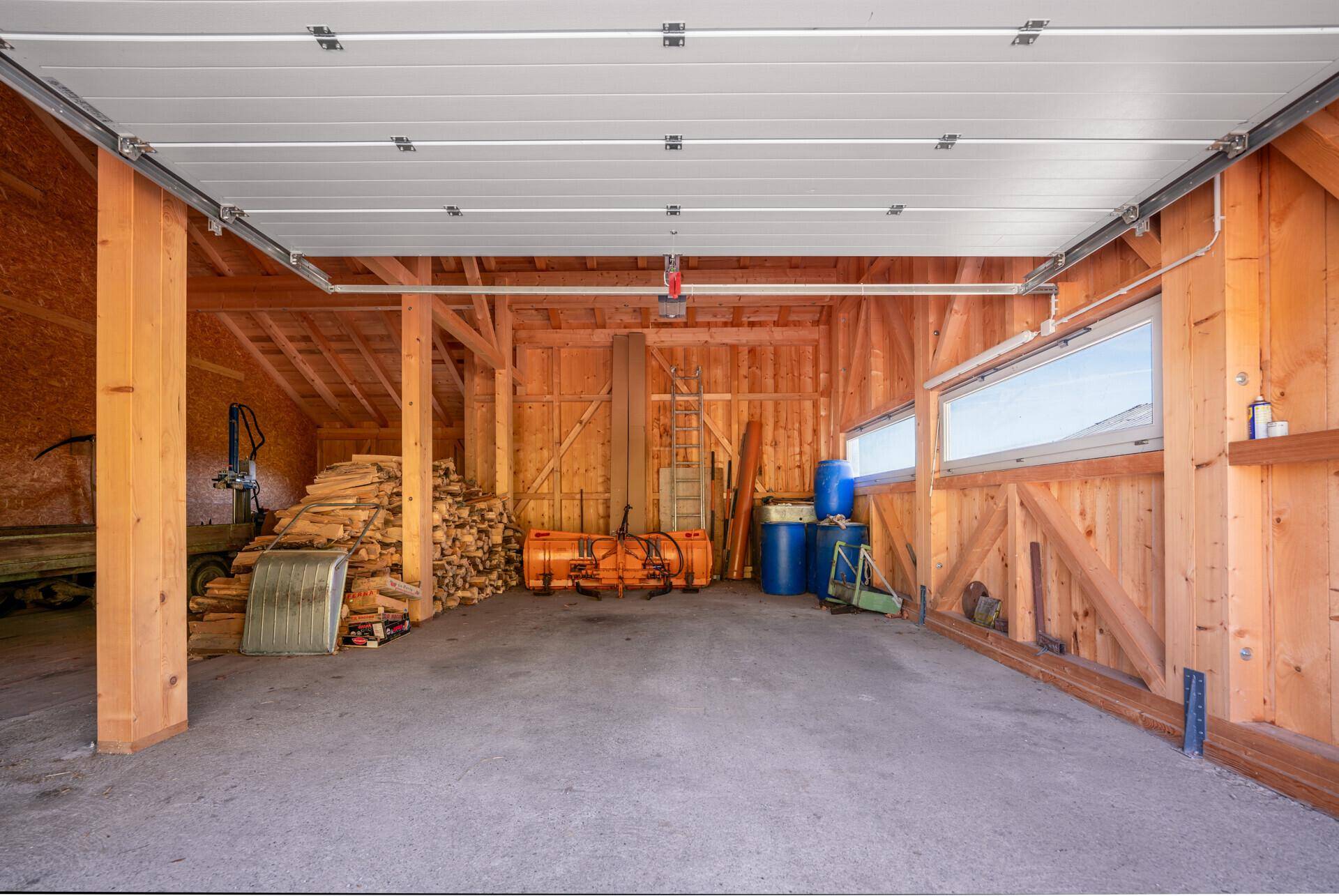 Geräumige Garage mit offenem Tor, Holzkonstruktion, Betonboden und gestapeltem Brennholz.