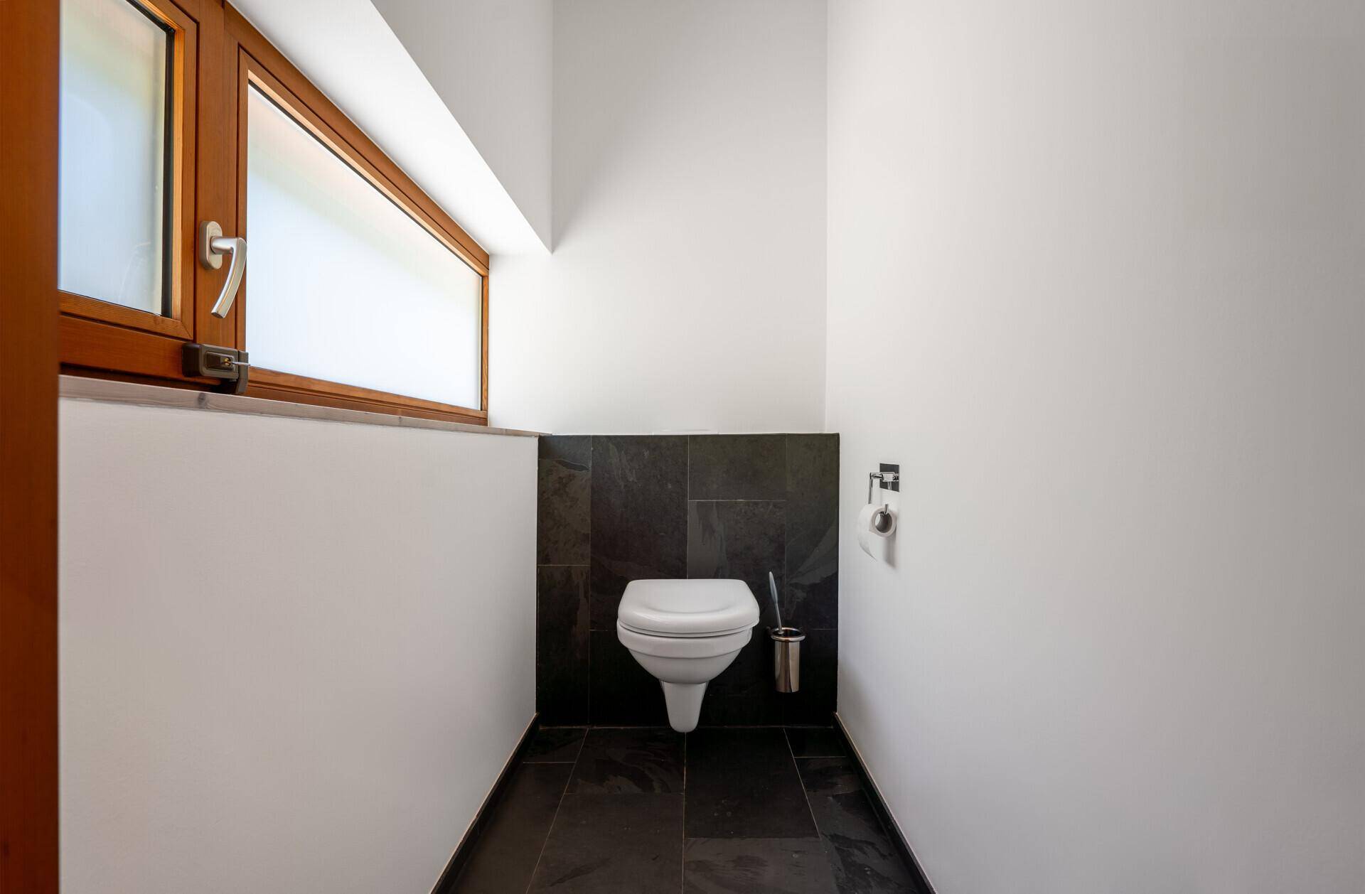 Separate Toilette mit Wand-WC, dunklen Fliesen und kleinem Fenster.