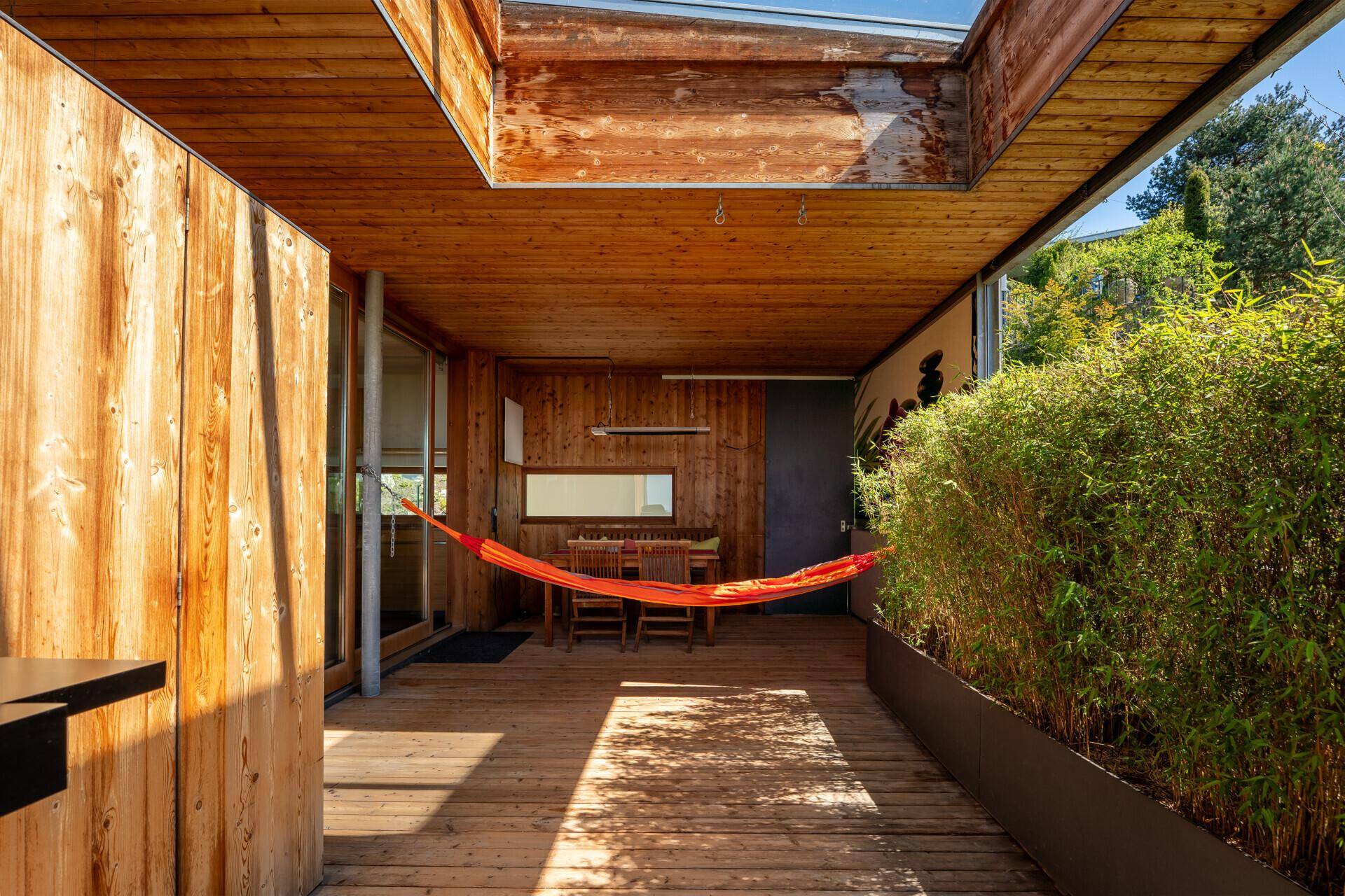 Überdachte Holzterrasse mit Hängematte, Essbereich und üppiger Bepflanzung.