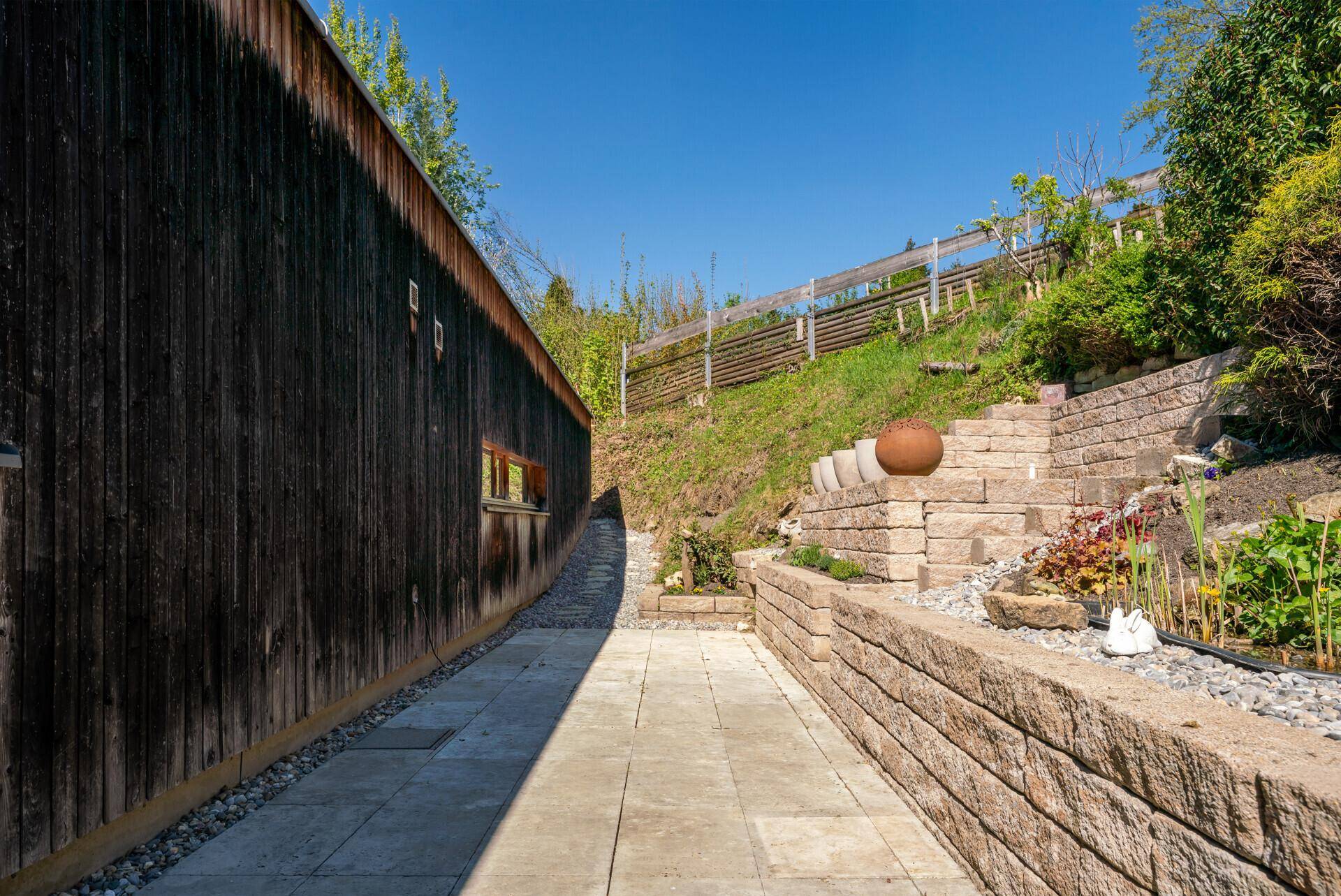 Gepflasterter Gartenweg entlang einer Holzwand mit terrassierten Beeten und Steinmauern.