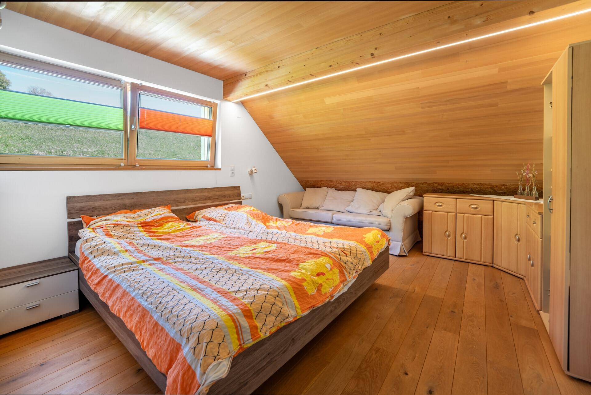Schlafzimmer mit Doppelbett, Holzdielenboden und Holzschrägdecke, helle Fenster mit farbigen Rollos.