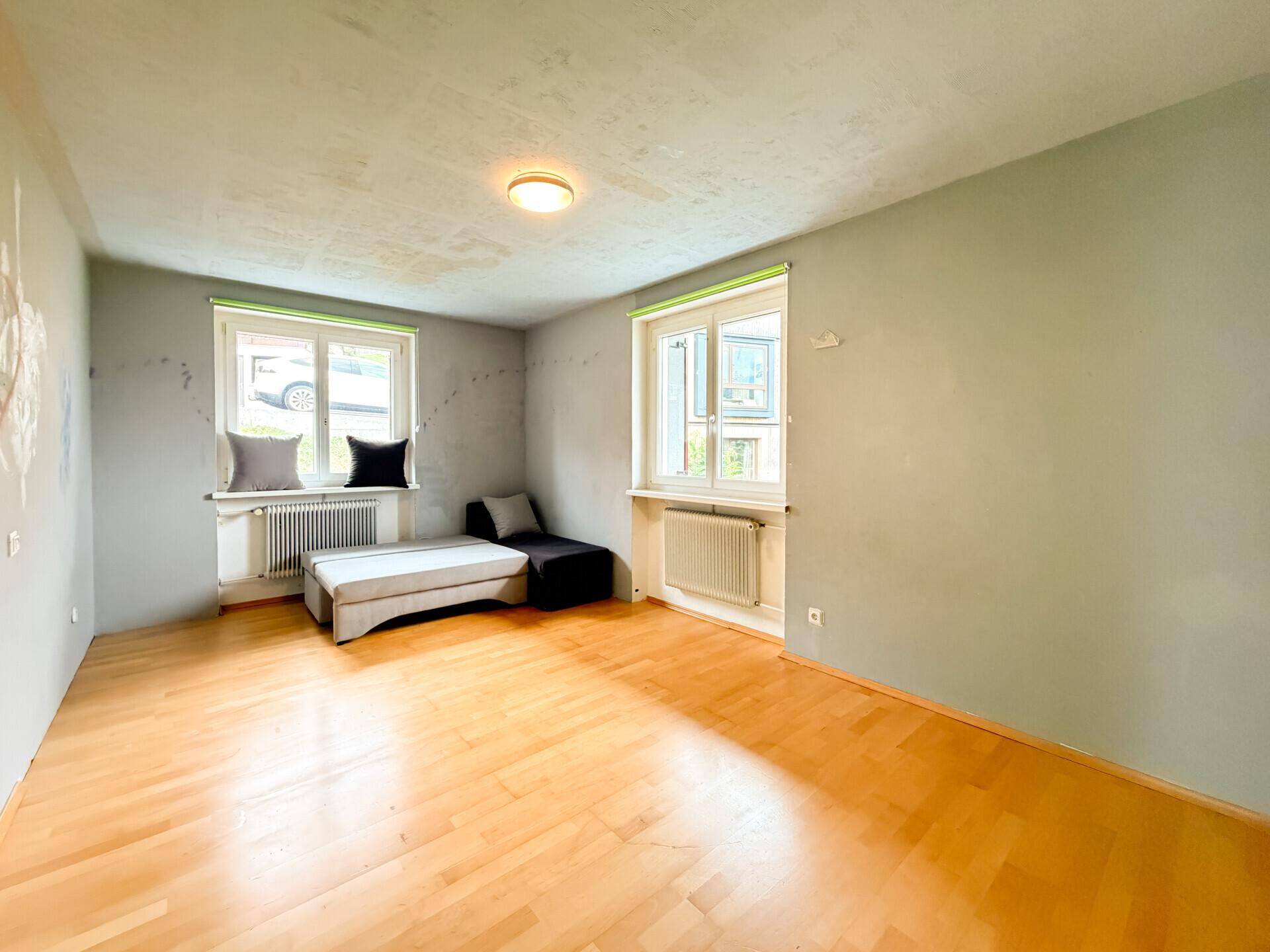Freundliches Zimmer mit Holzboden und zwei Fenstern, die viel Tageslicht hereinlassen.