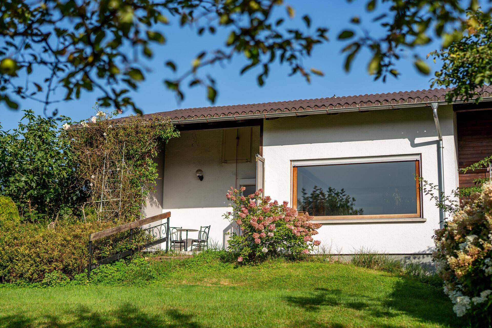 Hausfassade mit kleiner Terrasse und Sitzgelegenheit, umgeben von Grün.