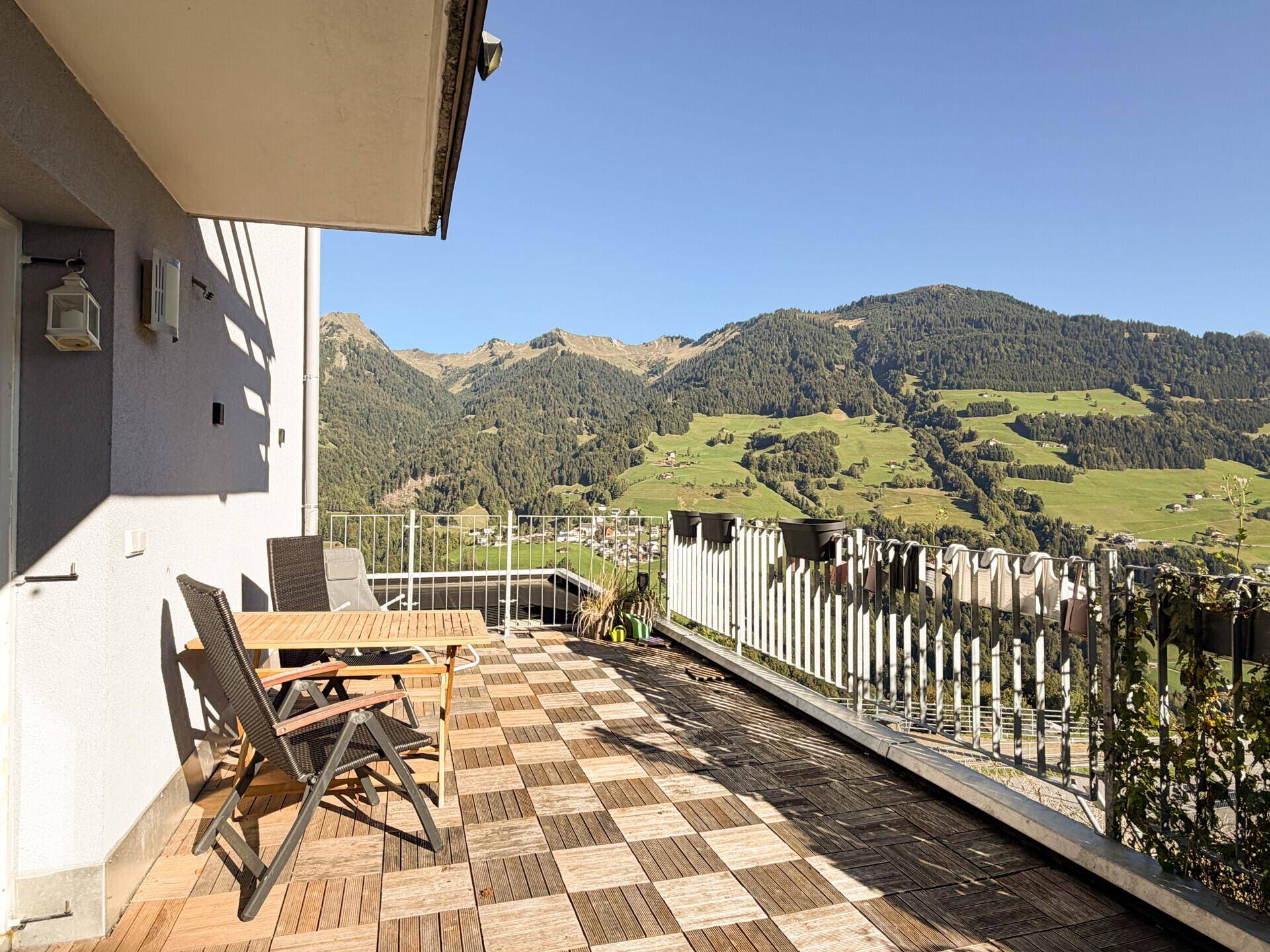 Geräumiger Balkon mit Holzboden und Sitzgelegenheiten, bietet einen weiten Blick auf die Berge und das Tal.
