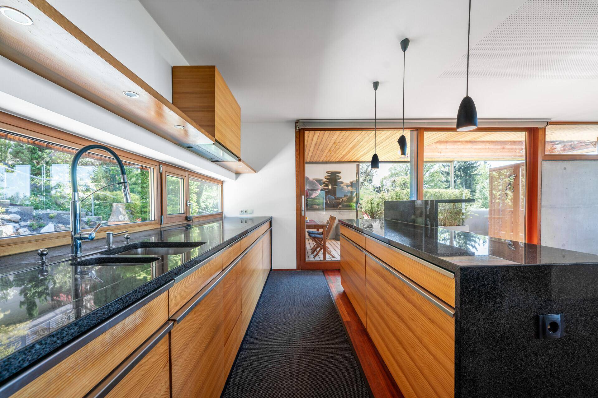 Lange Küchenzeile mit Holzschränken, dunklen Arbeitsplatten und Fensterfront zum Garten.