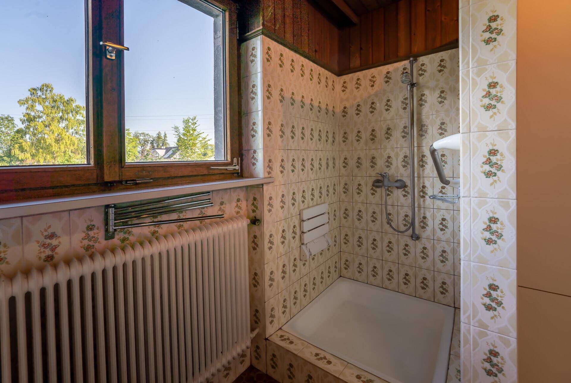 Duschbereich im Badezimmer mit gemusterten Fliesen, Fenster und Heizkörper.