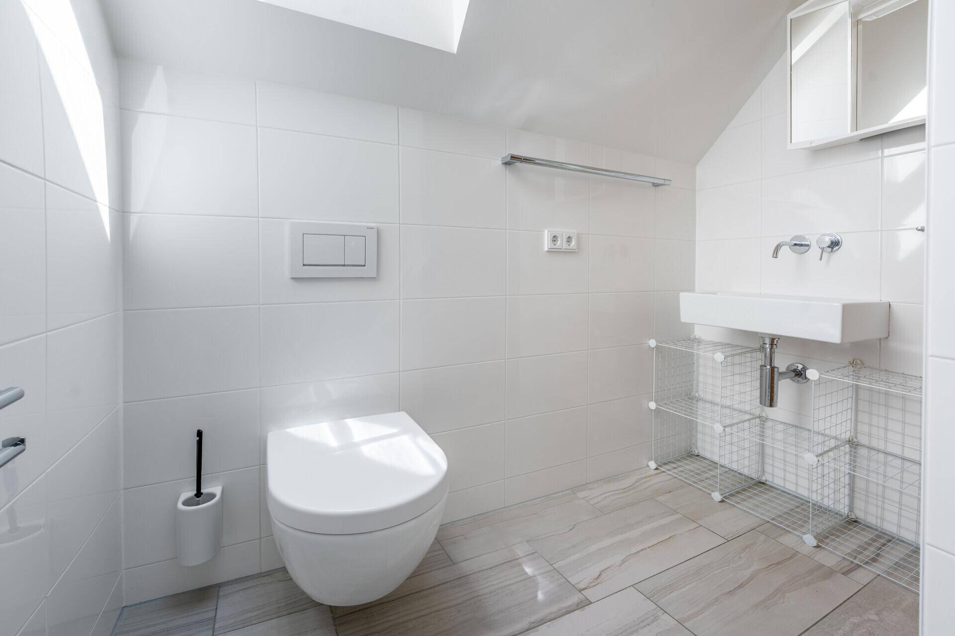 Modernes Badezimmer mit wandhängendem WC, weißen Fliesen und einem kleinen Waschbeckenbereich.