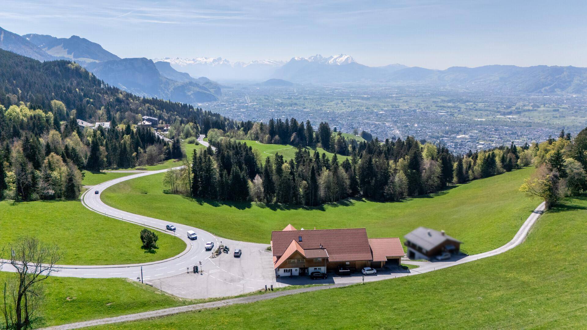 Panoramablick auf ein Gebäudeensemble in grüner Berglandschaft mit einer Stadt im Hintergrund.