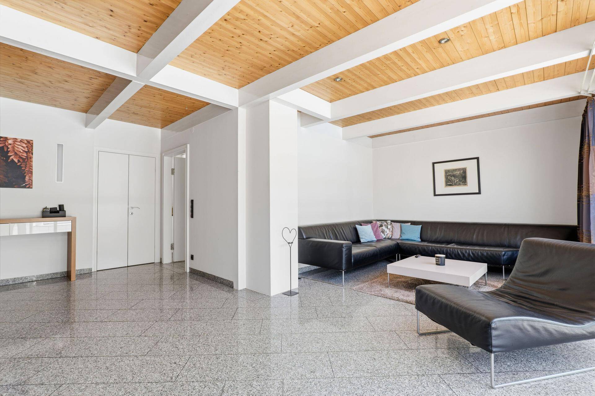 Geräumiges Wohnzimmer mit dunklem Ecksofa, Holzbalkendecke und Steinboden.