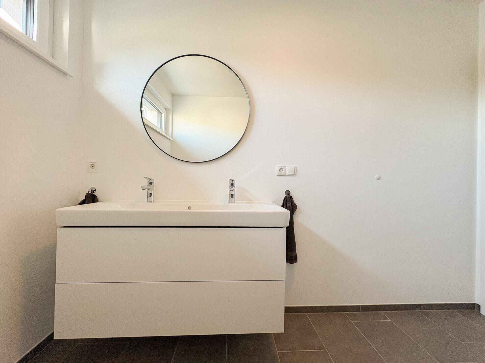 Modernes Badezimmer mit Doppelwaschbecken, rundem Spiegel und großem Fenster.