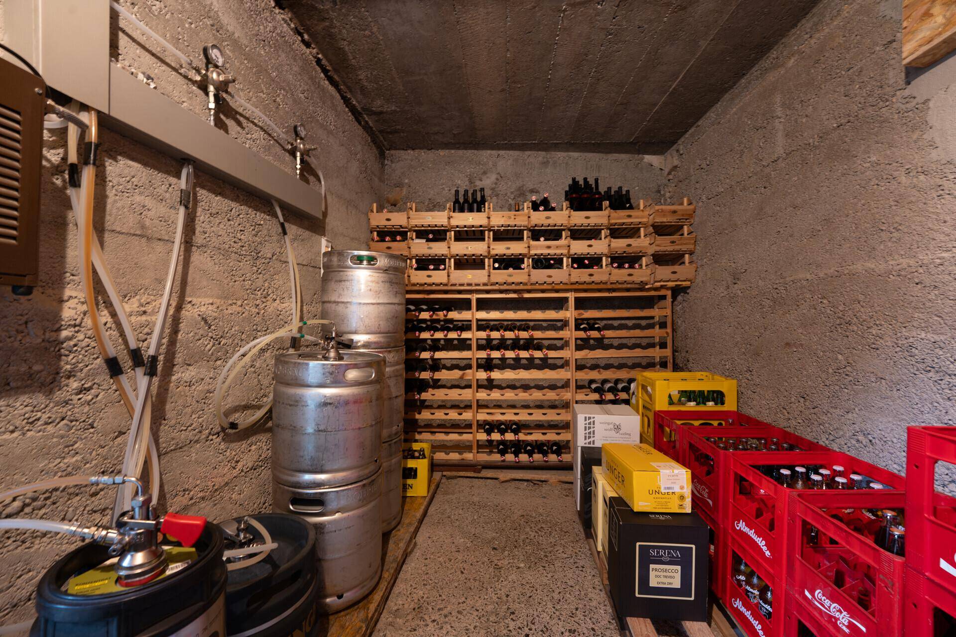 Kellerraum mit Betonwänden, Bierfässern und Holzregalen, gefüllt mit Weinflaschen für Lagerung.