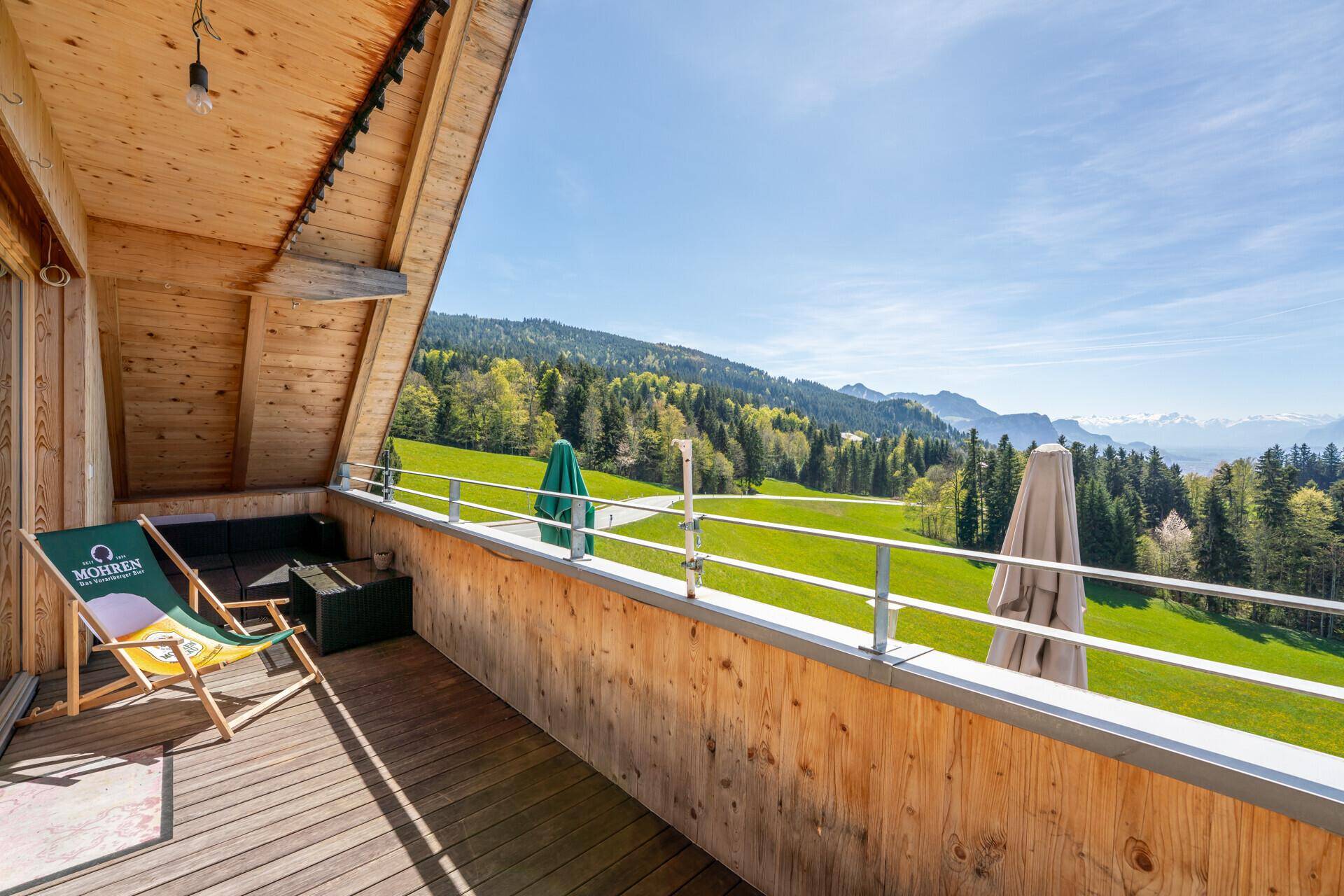 Geräumiger Holzbalkon mit Liegestuhl und Sonnenschirmen, bietet einen weiten Blick ins Grüne und auf Berge.