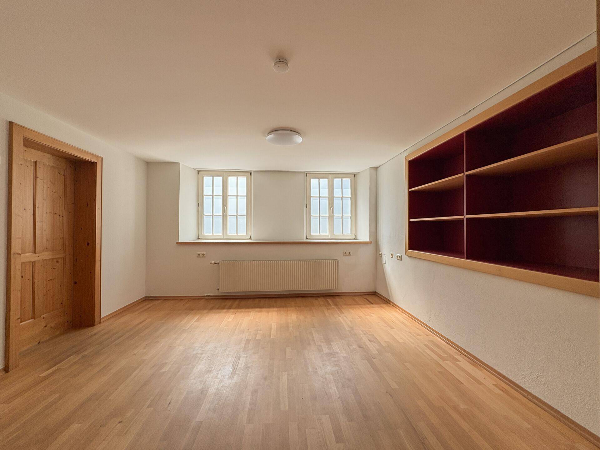 Geräumiger Raum mit Holzparkett, zwei Fenstern und eingebauten Regalen.