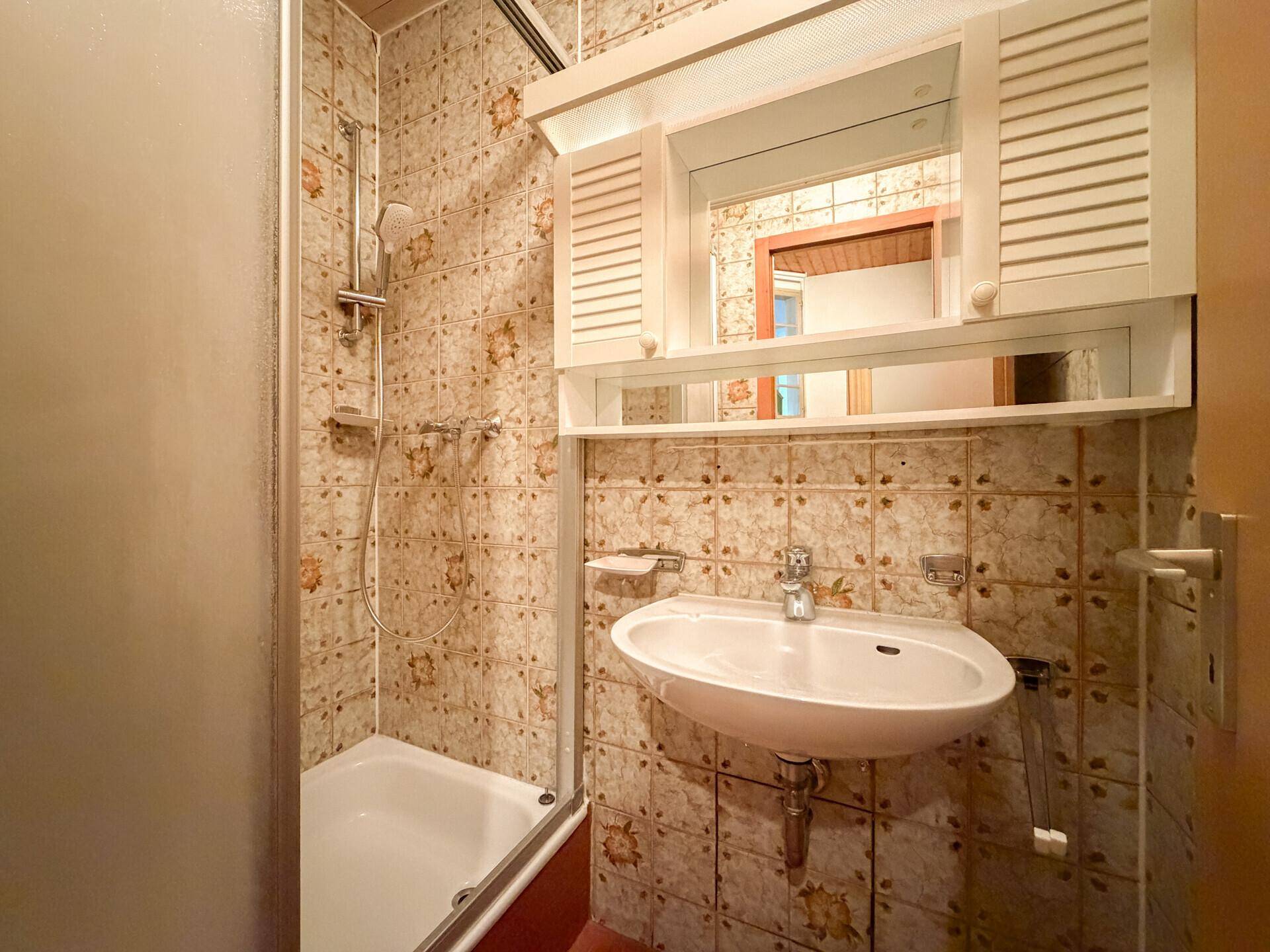 Kleines Badezimmer mit Dusche, Waschbecken und gemusterten Fliesen im 80er-Jahre-Stil.