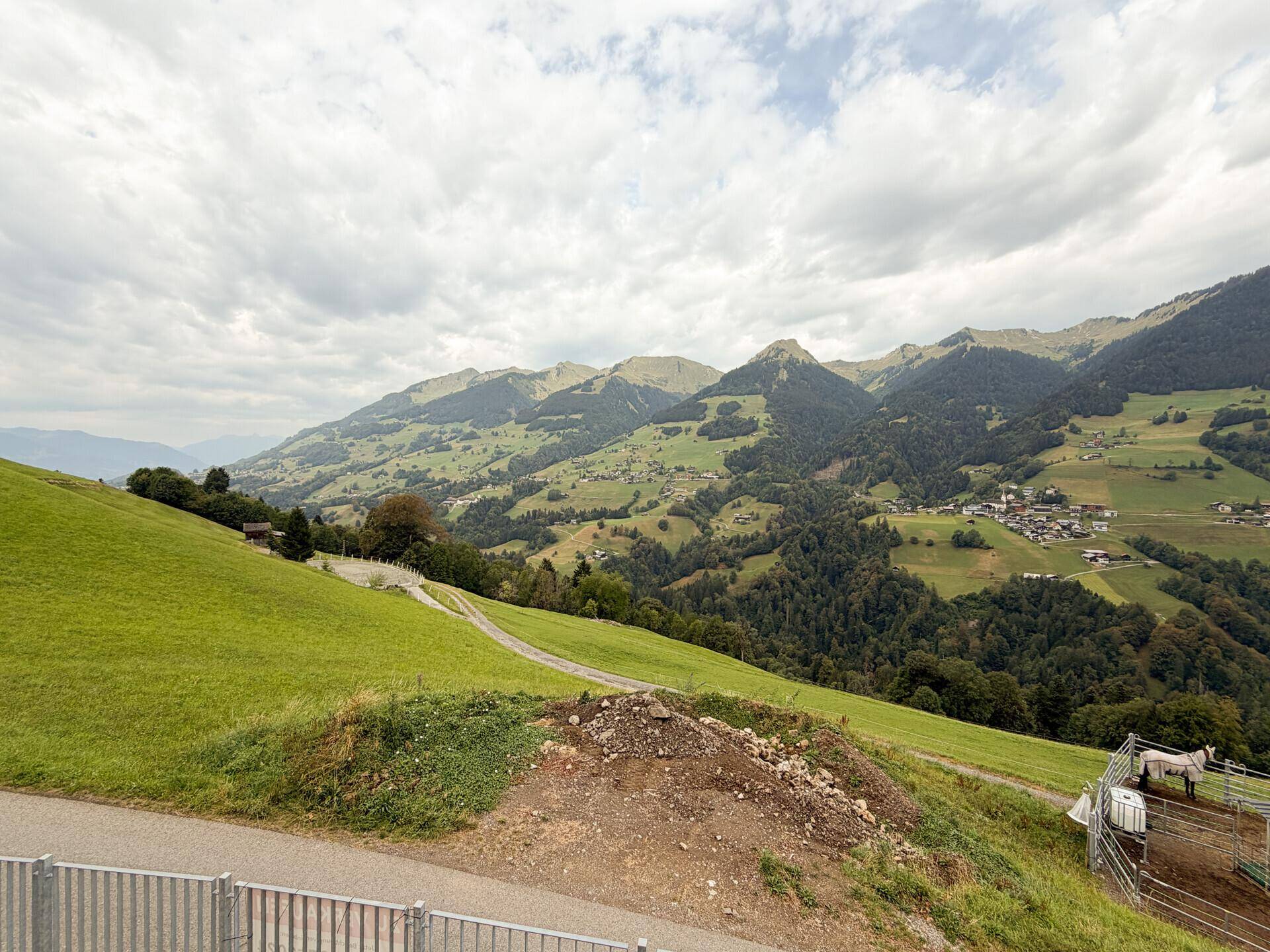 Malerischer Ausblick auf weite grüne Hügel und majestätische Berge unter bewölktem Himmel.
