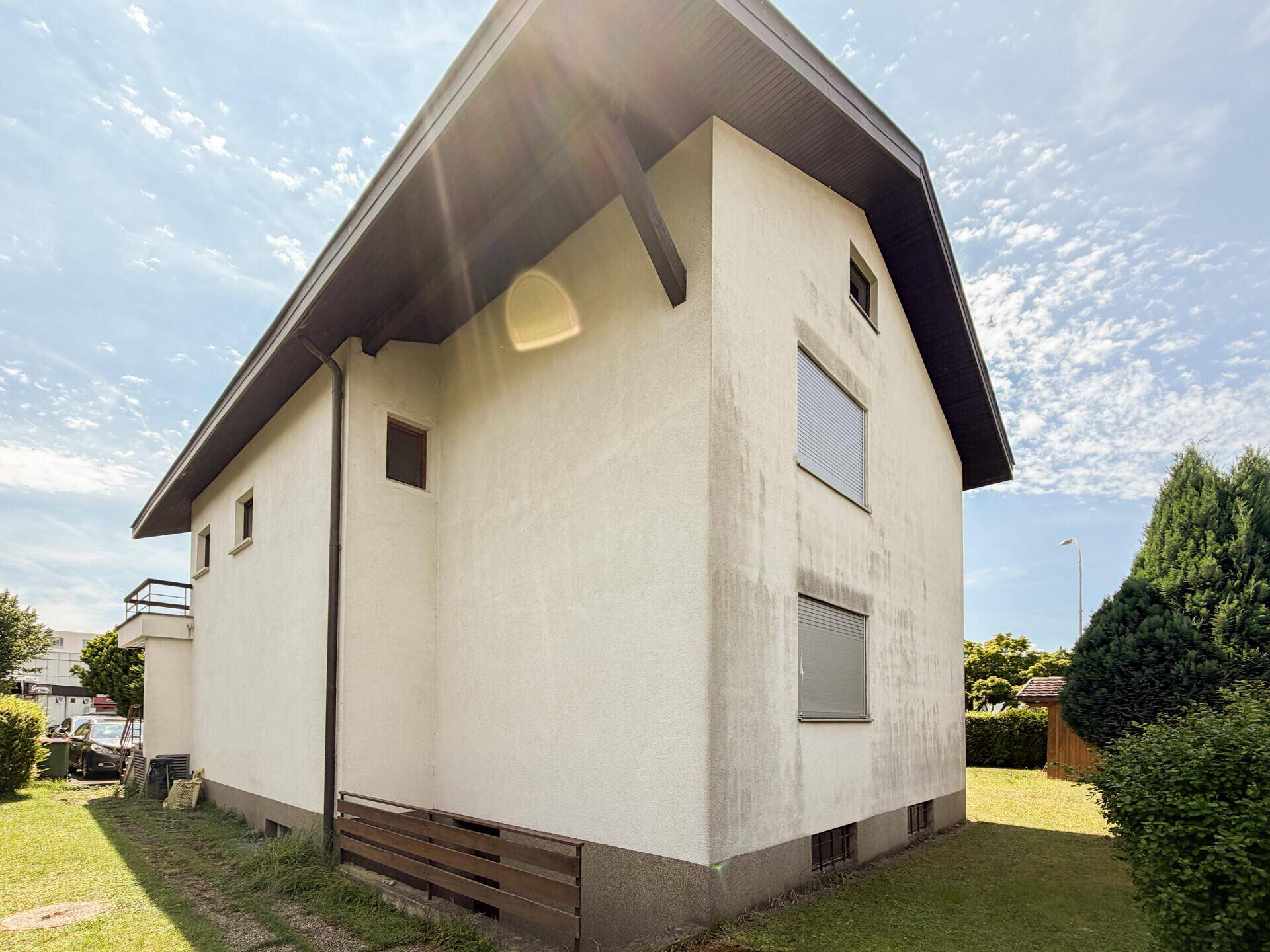 Schräge Außenansicht des Hauses mit weißer Fassade, dunklem Dachüberstand und Fenstern mit Rollläden.