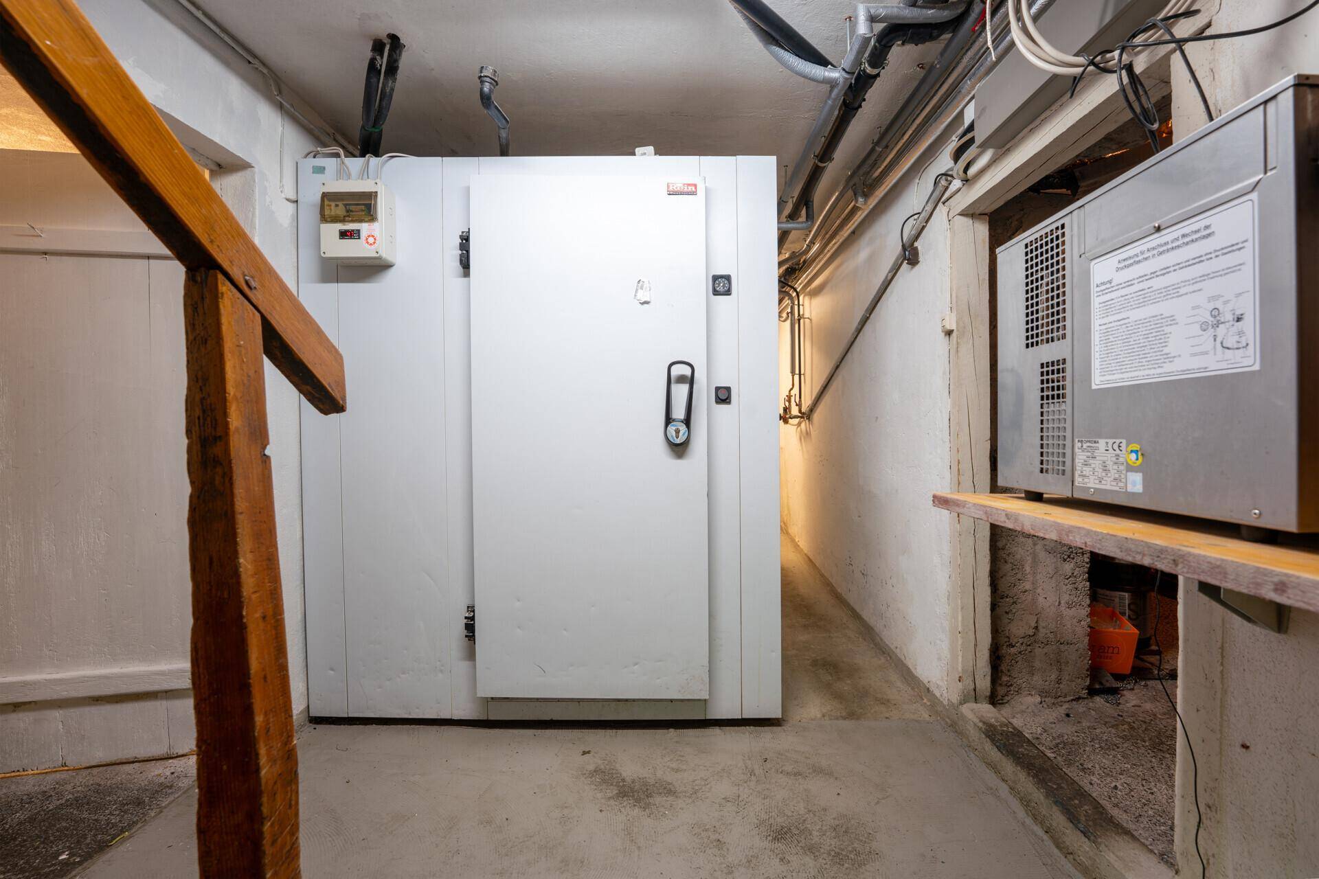 Kühlraum mit großer weißer Tür, Betonboden und sichtbaren Leitungen an der Decke für Lagerzwecke.