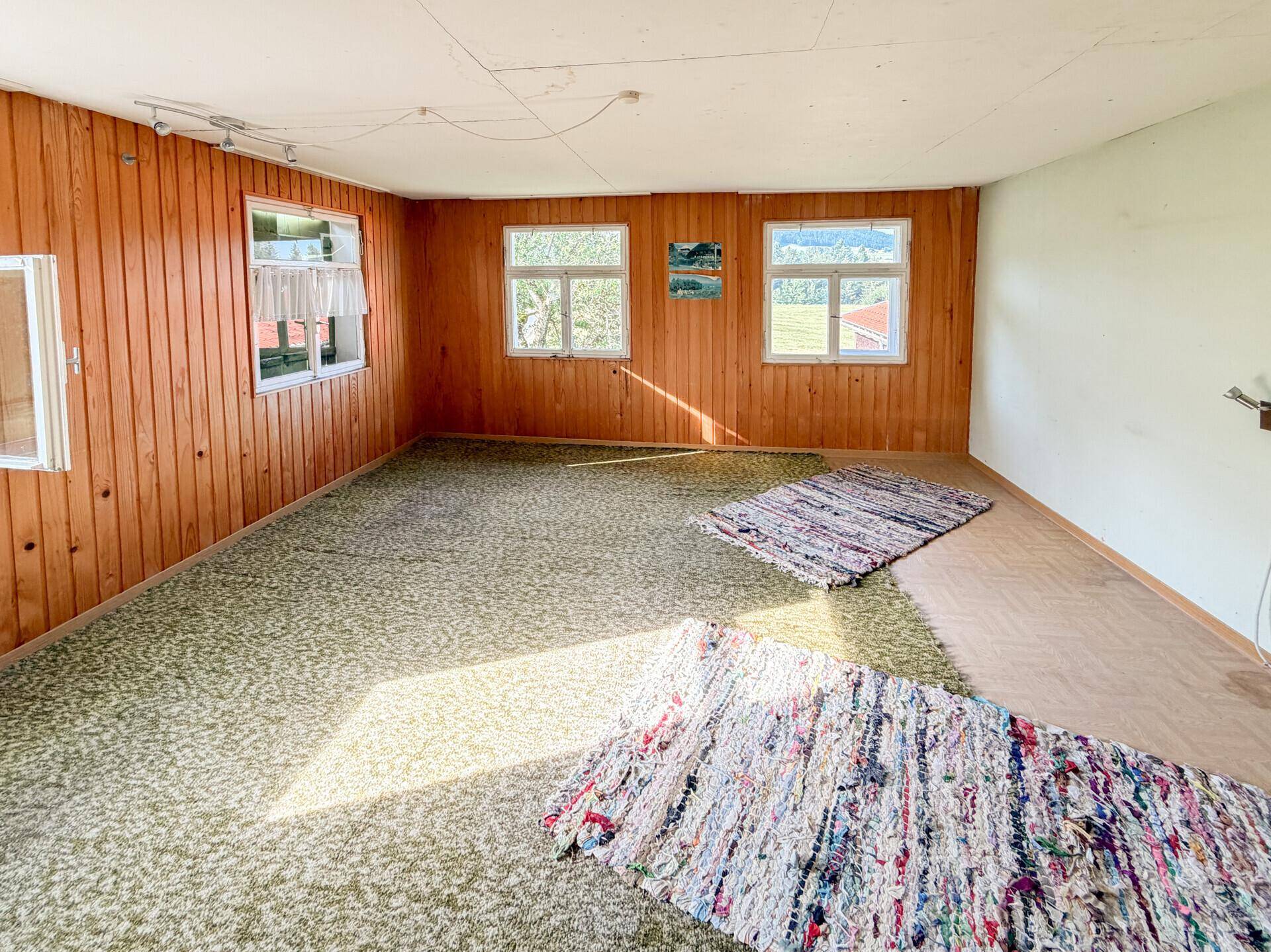 Geräumiger Raum mit Holzwänden, grünem Teppichboden und mehreren Fenstern für viel Tageslicht.
