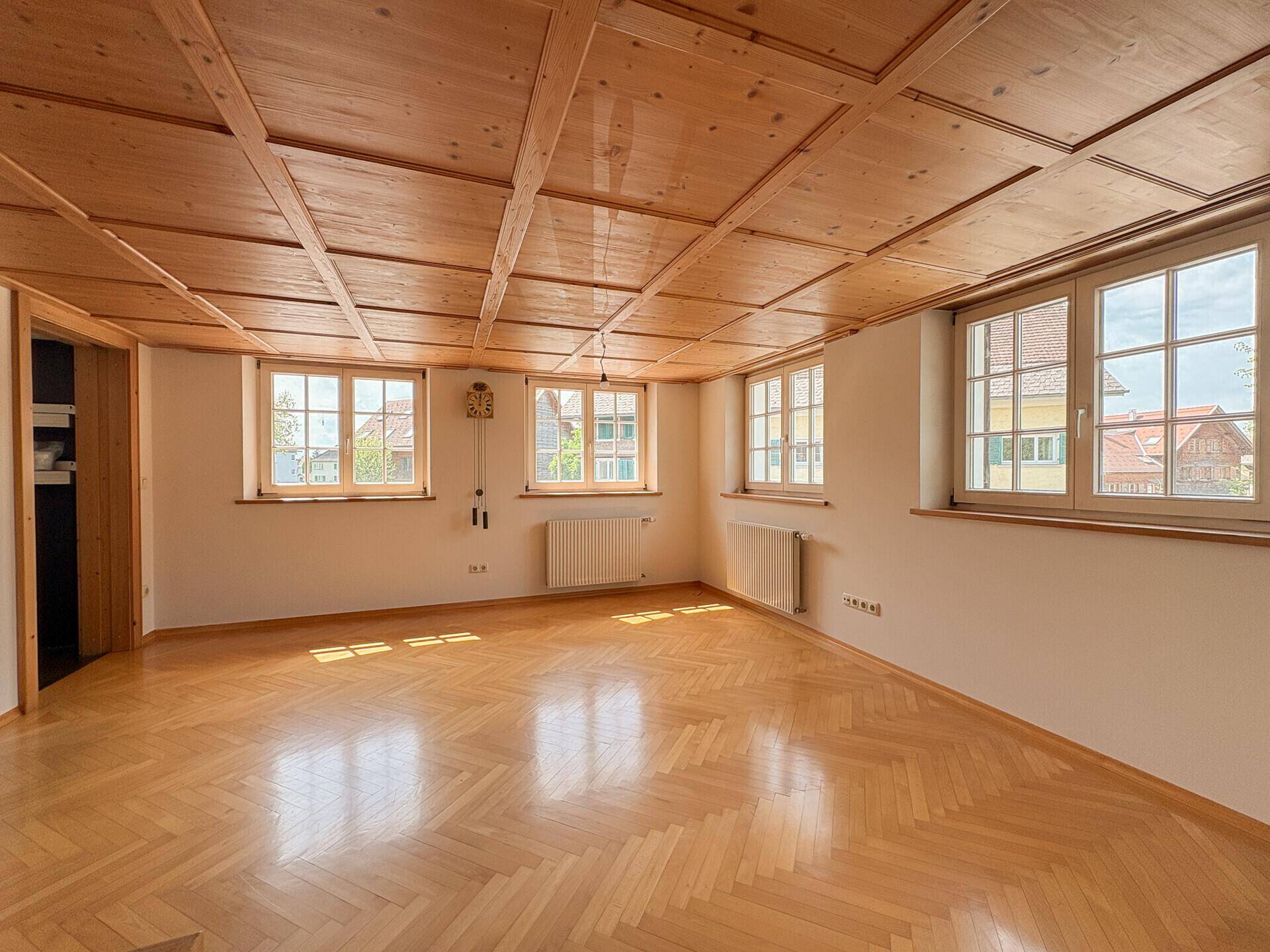 Helles Wohnzimmer mit Holzbalkendecke, Fischgrätparkett und mehreren Fenstern.