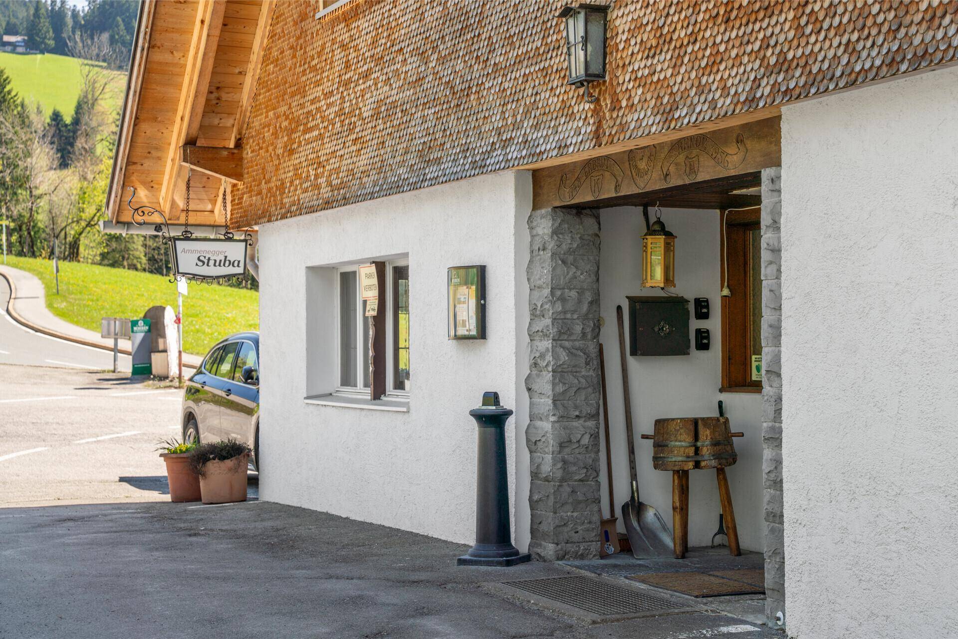 Eingangsbereich eines traditionellen Gebäudes mit Holzelementen und einem Schild, umgeben von ländlicher Landschaft.