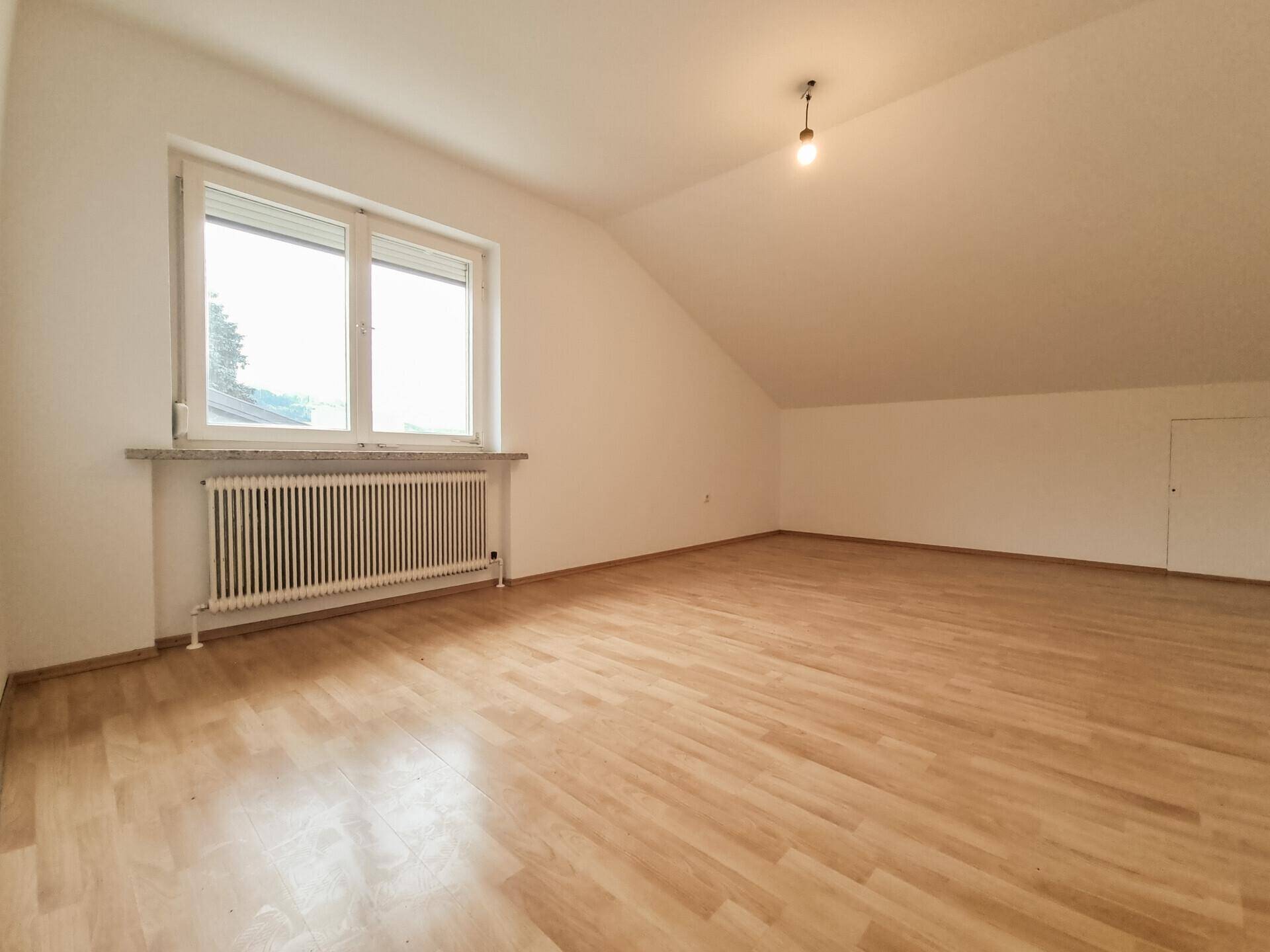 Heller, leerer Raum mit großem Fenster, Heizkörper und Dachschräge.