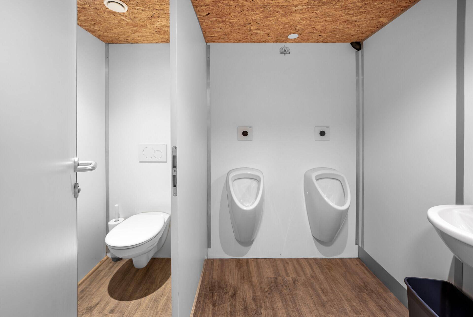 Herren-WC mit zwei Urinalen, einer Wandtoilette und einem Waschbecken, ausgestattet mit Holzoptikboden.