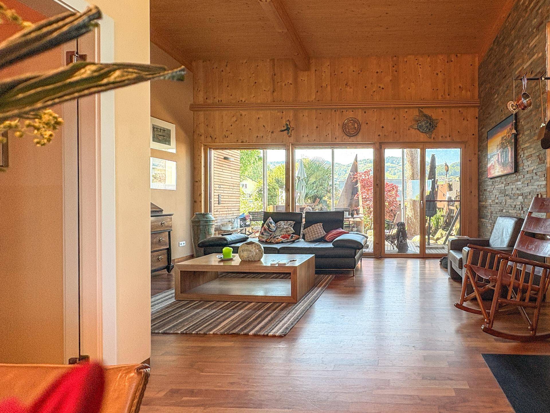 Gemütliches Wohnzimmer mit Holzwänden, Holzboden und großen Fenstern mit Blick ins Grüne.