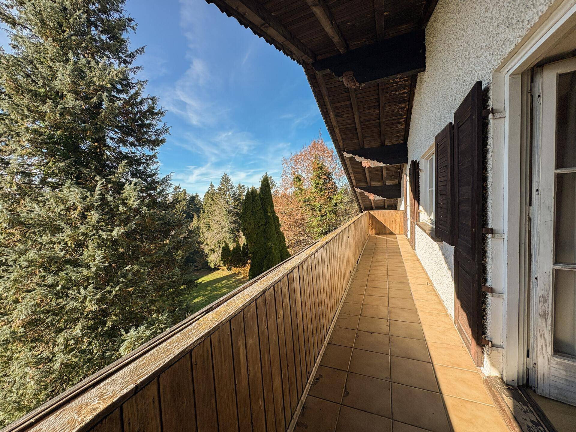 Langer Balkon mit Fliesenboden und Holzgeländer, umgeben von Bäumen.
