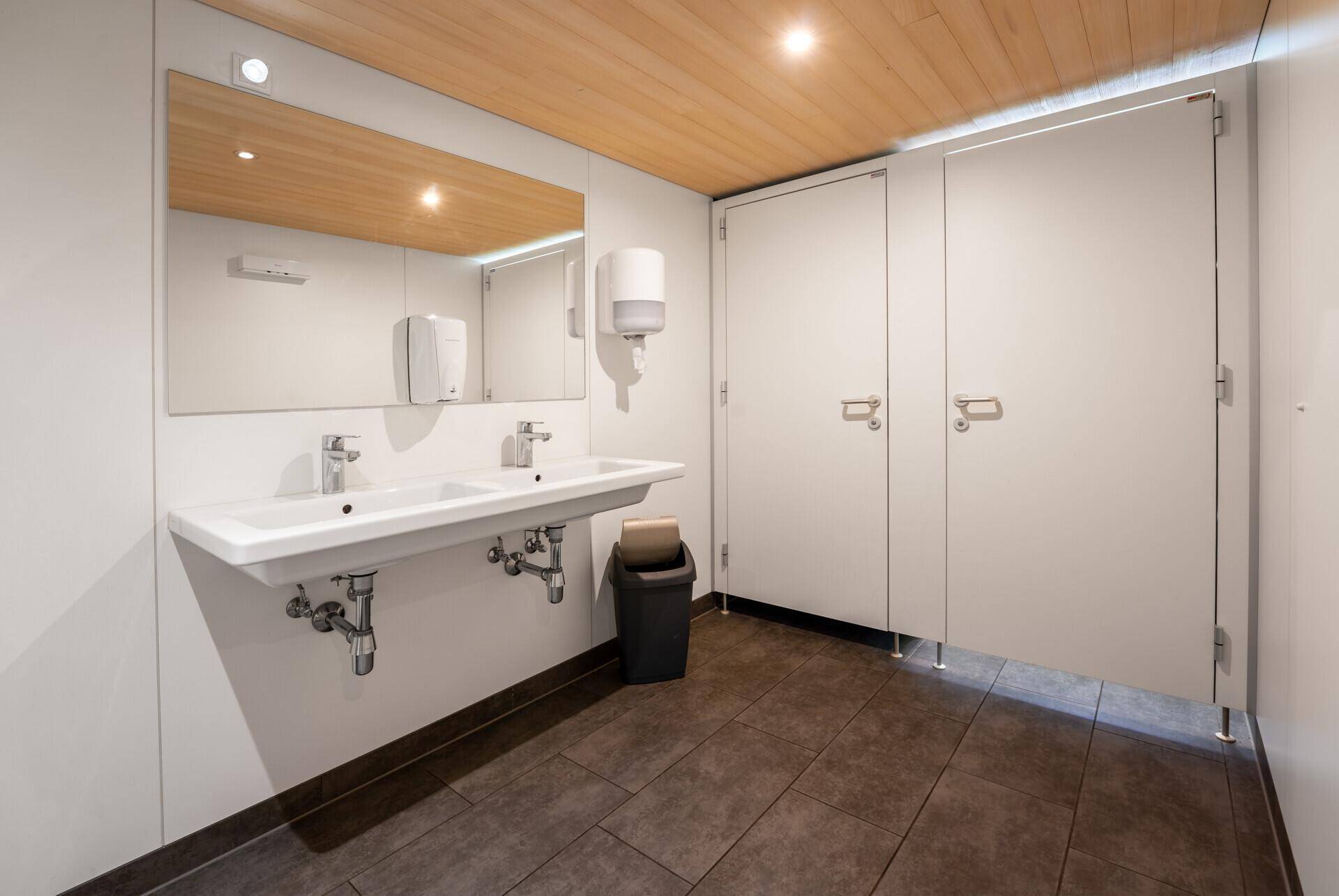 Moderner und sauberer Waschraum mit zwei Waschbecken, großem Spiegel und Holzakzenten an der Decke.