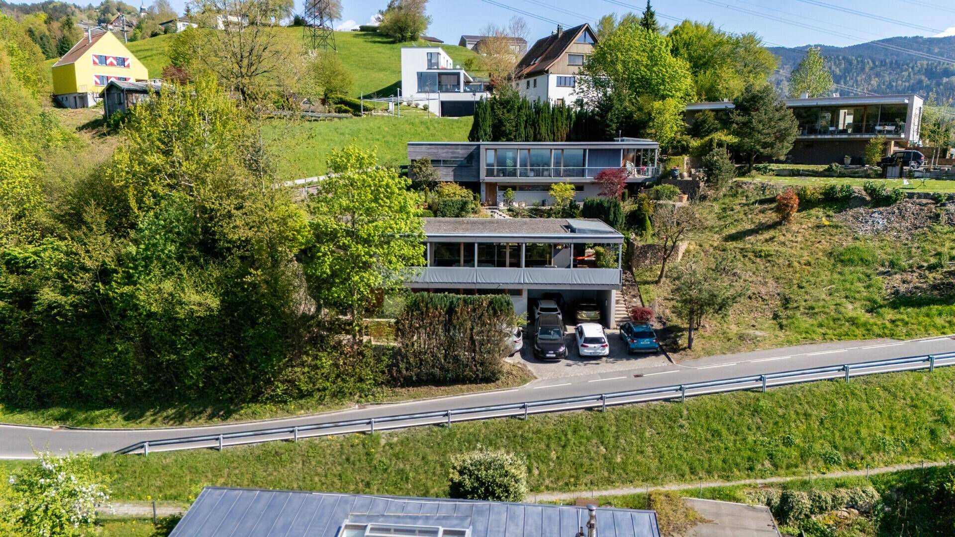 Luftaufnahme des modernen Hauses mit Flachdach und umliegenden Gebäuden in grüner Hanglage.
