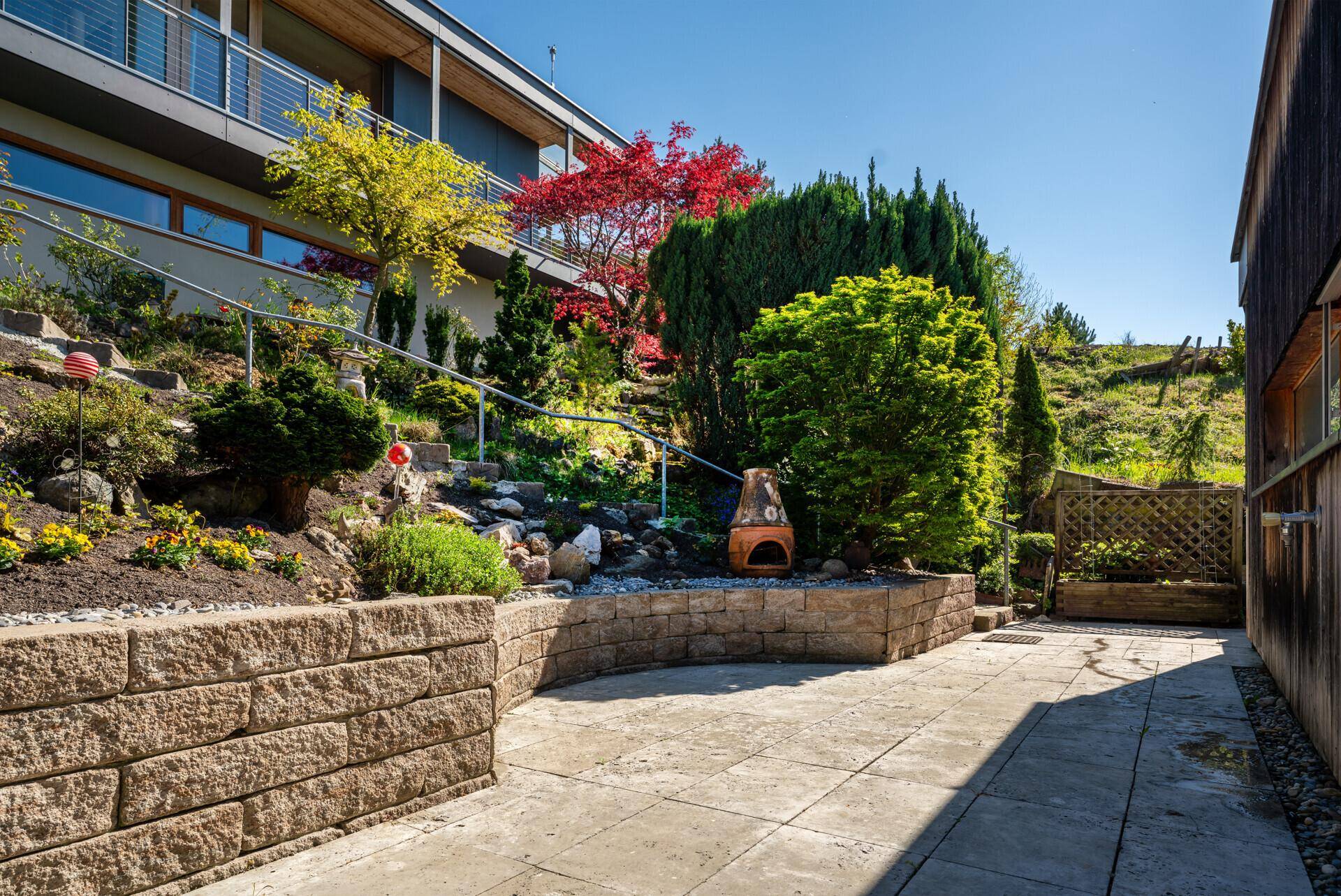 Terrassierter Gartenbereich mit Steinmauern, Pflanzen und einem Außenkamin.